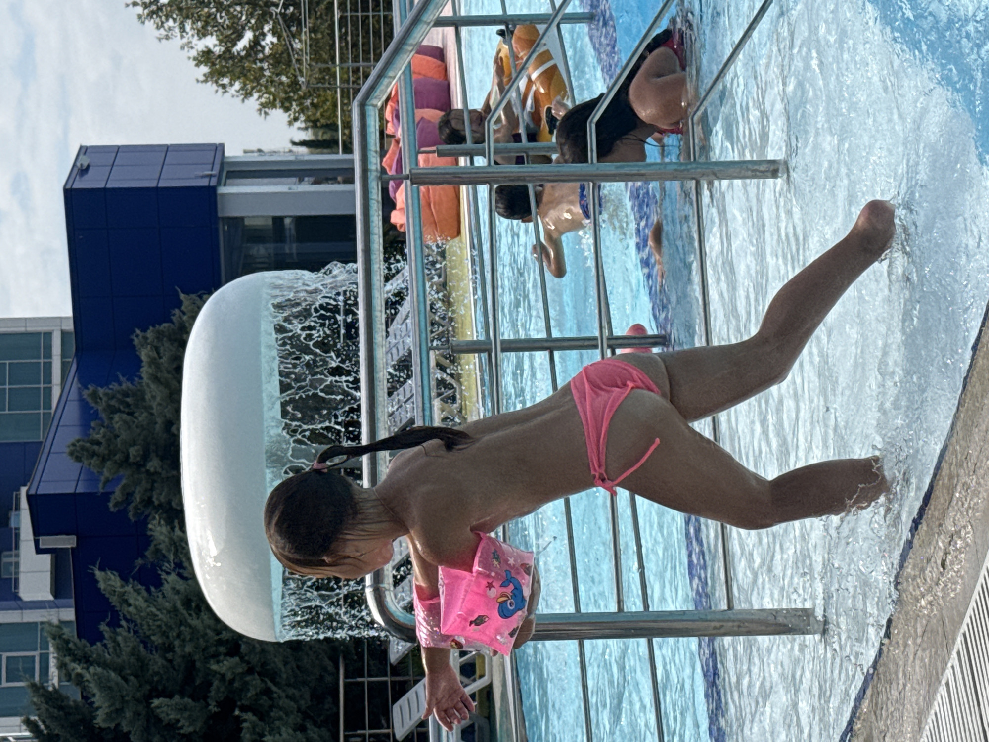 In Sevastopol, Ukraine, a young girl in a pink bikini and arm floaties is captured in motion at a pool. She is leaning forward, her body angled towards the water, with her right arm extended, splashing. The pool water is bright blue and filled with ripples and splashes, suggesting active play. Behind her, a large, smooth, white structure appears to be part of a water feature, with water cascading over its edge. In the background, a modern blue building with geometric window patterns and some greenery are visible. Other people are present in the pool area, including a person in orange floatation devices and another child partially submerged in the water. The lighting suggests it is daytime, likely sunny given the bright water and shadows. The overall scene conveys a sense of summer fun and aquatic activity in an outdoor recreational setting.