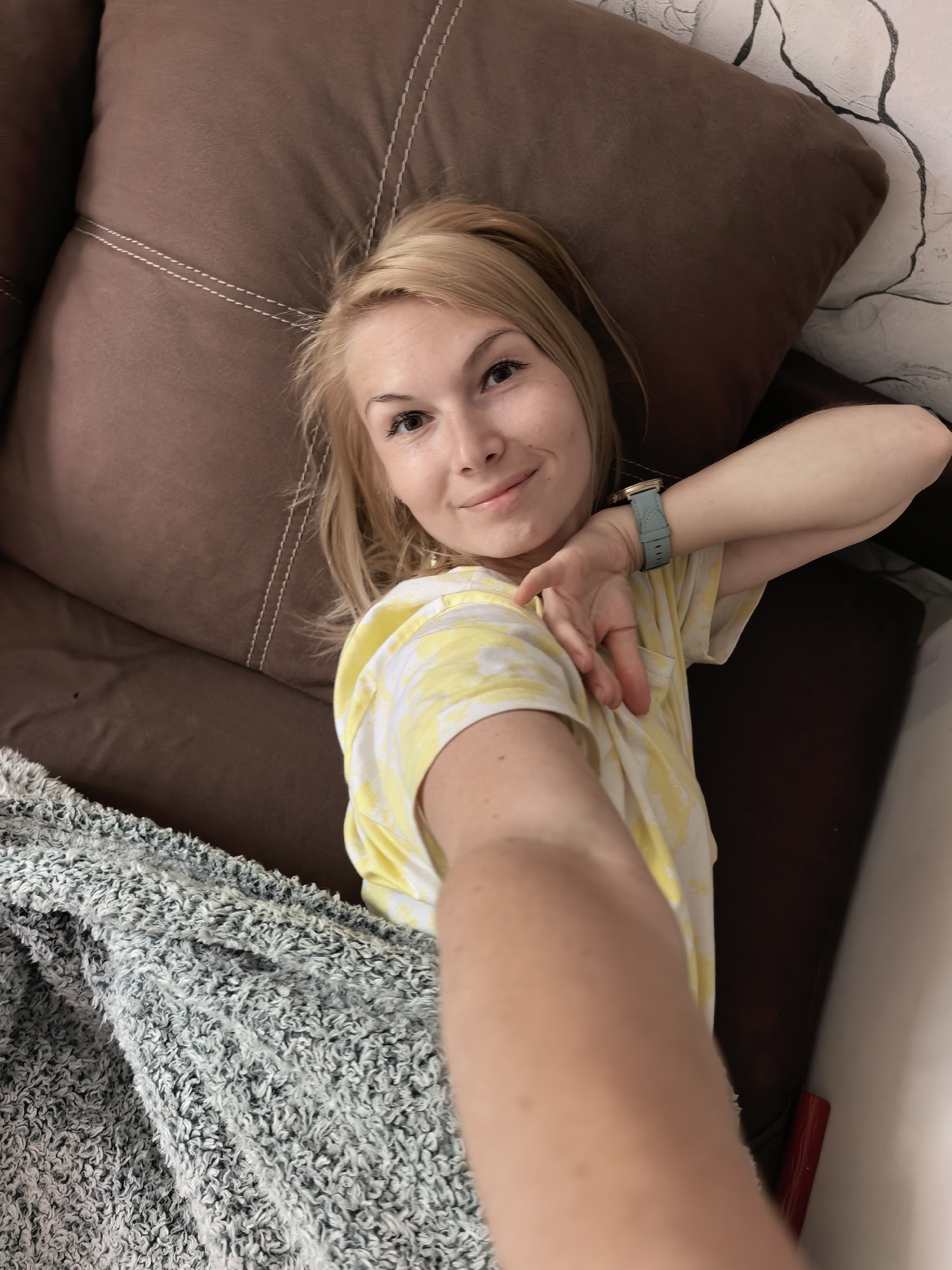 A woman is reclining on a brown couch covered partially by a grey and green blanket. She is blonde with fair skin and is wearing a yellow and white tie-dye shirt and a blue smartwatch. She is holding her arm out in a selfie-style pose. The location is likely indoors, with a wall visible behind the couch. There is no information to indicate the city of Sterlitamak, Russia.