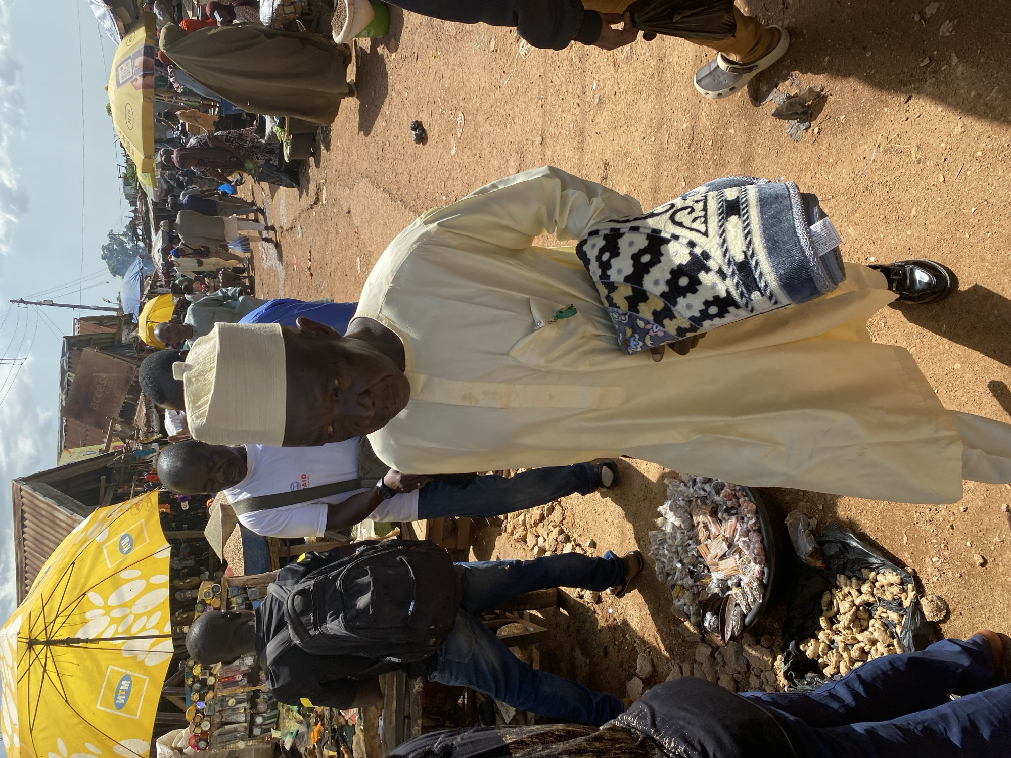The image captures a lively market scene in Bukuru, Nigeria. The central subject is a man wearing a light yellow kaftan and a white cap, holding a folded blanket with a patterned design. He is standing on a dirt path, surrounded by other people and market stalls. To his left, a bright yellow umbrella with "MTN" branding stands out, suggesting a commercial area. In the foreground, another man is visible, wearing a black backpack and blue jeans, with his legs crossed. To the right of the man in the kaftan, there are two bowls of goods, one containing various small items wrapped in plastic and the other filled with ginger roots in a black plastic bag. The ground is covered in dry, reddish-brown dirt, with some scattered stones and debris.
The atmosphere appears busy and active, typical of a market day. The lighting suggests it is daytime, likely mid-morning or afternoon, with shadows indicating the sun's position. The expressions on the faces of the people are not clearly discernible due to the image's orientation and resolution, but the overall impression is one of everyday commerce. The background shows more stalls and people, hinting at the scale of the market. The clothing styles of the individuals, particularly the kaftan and cap, are indicative of the region. The presence of various goods for sale, from textiles (the blanket) to produce (ginger) and small packaged items, highlights the diversity of products offered in the market. The image provides a glimpse into the vibrant daily life and economic activity of a Nigerian marketplace.