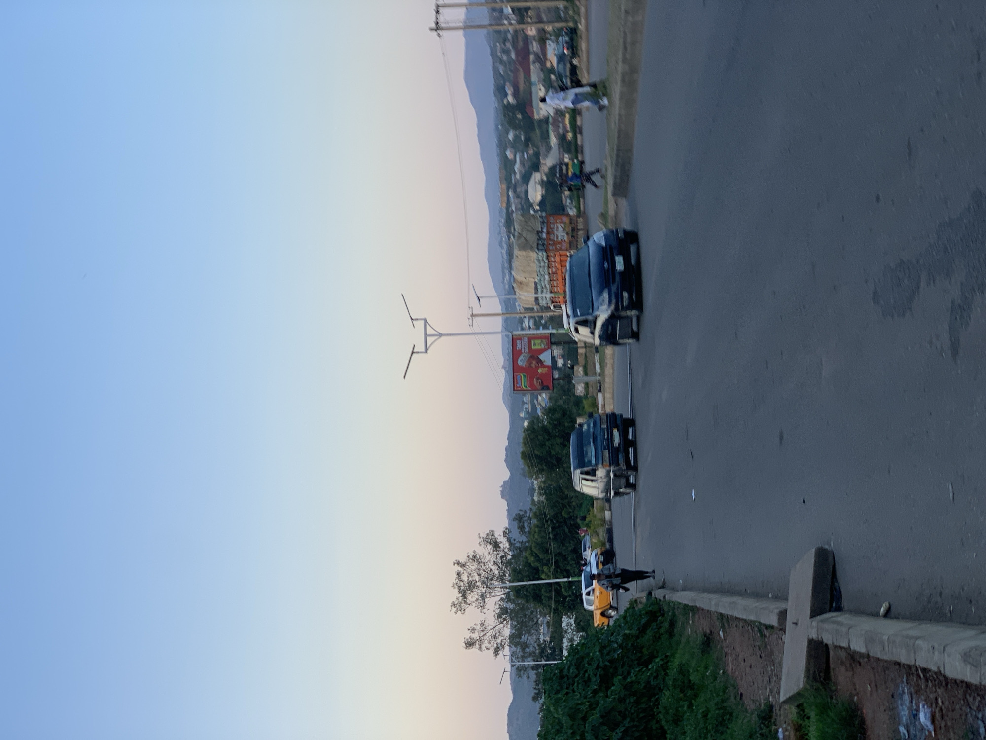 The image depicts a street scene in Jos, Nigeria, during the late afternoon or early evening. The sky is a clear gradient of light blue fading to pale yellow towards the horizon. In the foreground, a grey asphalt road is visible with a concrete curb on the left side, lined with overgrown green vegetation and some litter. A concrete slab has been dislodged from the curb, creating an uneven surface.
Several vehicles are present on the road. A dark blue van is moving away from the camera, slightly to the right of center. To the left, a light yellow pickup truck is parked, with a dark SUV behind it. A person wearing dark clothing and carrying a bag is walking along the sidewalk on the left side of the road, heading away from the camera. Further down the road, another dark van is visible, as well as a few other cars and pedestrians.
In the middle ground, there are trees and buildings. A billboard with an advertisement is visible on the right side of the road. In the background, a range of hills forms the horizon, with scattered buildings and structures indicating a populated area. Streetlights are present along the road. The overall impression is one of a typical urban environment in Jos during a time of day with soft lighting.
