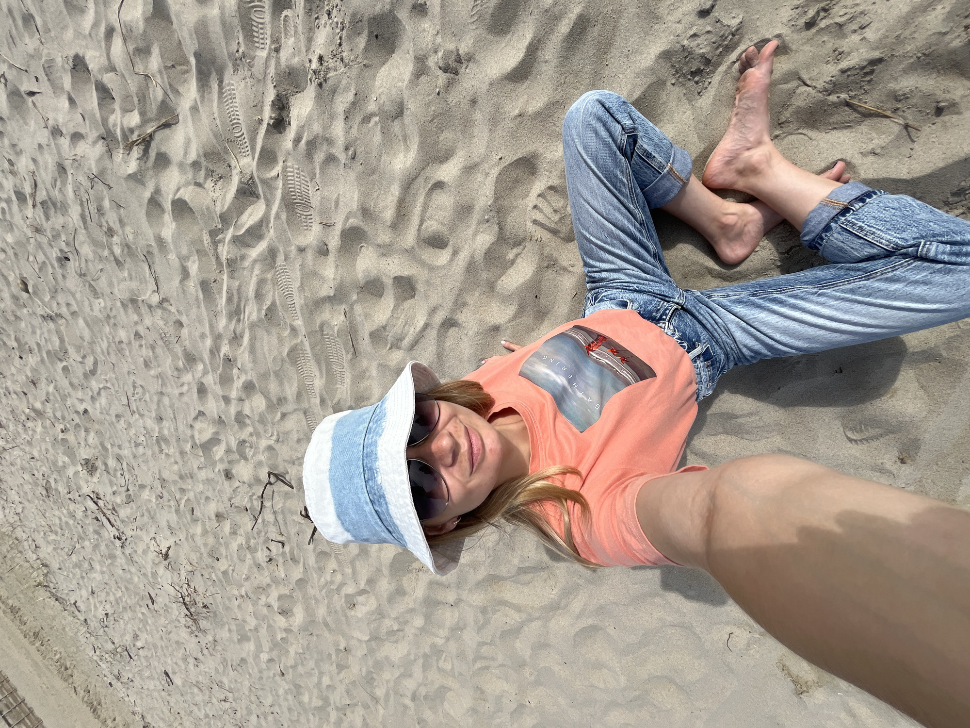 A woman is sitting on a sandy beach in Yantarny, Russia, taking a selfie. She is wearing a peach-colored t-shirt with a graphic print of a boat and text that reads "GATHERING," blue jeans, a white and blue bucket hat, and sunglasses. Her bare feet are resting on the sand. The beach is mostly bare with scattered driftwood and visible footprints in the sand. The sky is not visible in the frame.