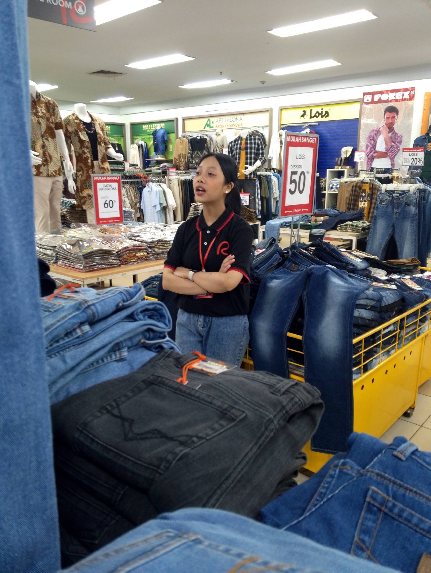 The image depicts the interior of a clothing store in Batam, Indonesia, likely a department store or a large retail chain. In the foreground, stacks of folded jeans, primarily denim blue and a dark charcoal grey, are displayed on a yellow retail fixture. A woman, an employee with dark hair and a black polo shirt with a red collar and a red lanyard, stands with her arms crossed, looking upwards and speaking. Behind her, various clothing racks are visible, displaying shirts and pants. Signs advertising discounts are prominently displayed, including a red sign with "60%" and another with "50%" for the brand "LOIS". A large poster featuring a male model wearing a plaid shirt and jeans for the brand "FOREVER" is also visible in the background, as are mannequins dressed in batik-patterned shirts. The lighting suggests an indoor commercial setting.