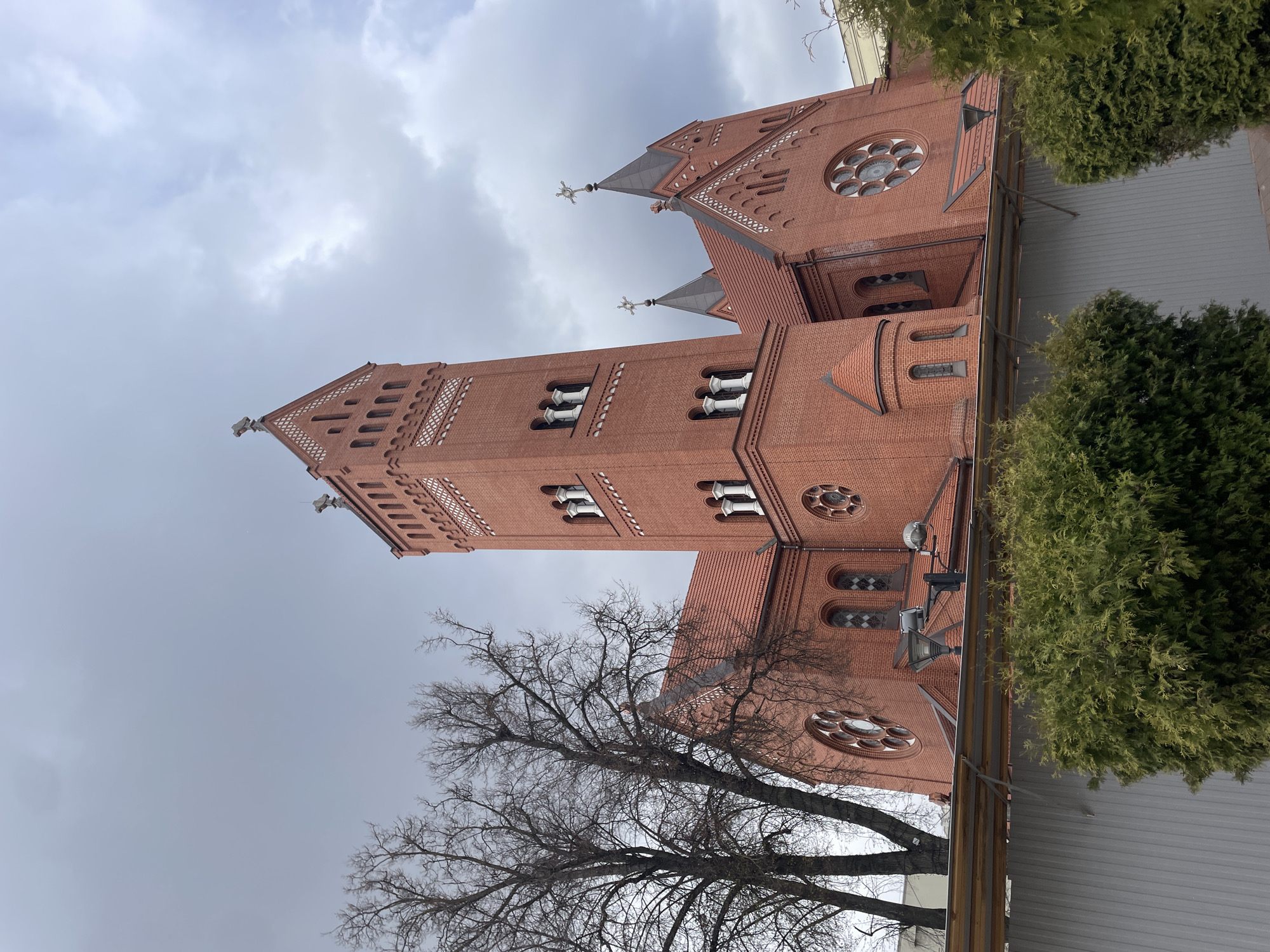 The image displays the exterior of a large, red-brick church under an overcast sky. The church exhibits prominent Gothic Revival architectural features, with a dominant central bell tower rising on the left side of the frame. This tower is adorned with multiple arched windows, intricate brick patterns, and culminates in a spire topped with a cross.
To the right of the main tower, another section of the church's facade is visible, featuring a large, ornate circular rose window. Two smaller spires, also crowned with crosses, emerge from the roofline in this area. The building's construction is characterized by its red brick, complemented by decorative lighter-colored brickwork in patterns and window surrounds. Various arched windows, some with clear panes and others with diamond-shaped detailing, punctuate the building's walls. Outdoor lighting fixtures are mounted on the church's exterior.
In the foreground and extending along the right side, several trees are present. A large deciduous tree with bare, intricate branches stands in the lower left portion of the image. To its right, dense evergreen shrubs or conifers provide a contrasting green, partially obscuring the lower parts of the church and a low fence or structure. The sky is uniformly grey and cloudy, indicating an overcast day. There are no visible people or discernible text in the photograph. This is likely the Church of Saints Simon and Helena (Red Church) in Minsk, Belarus.