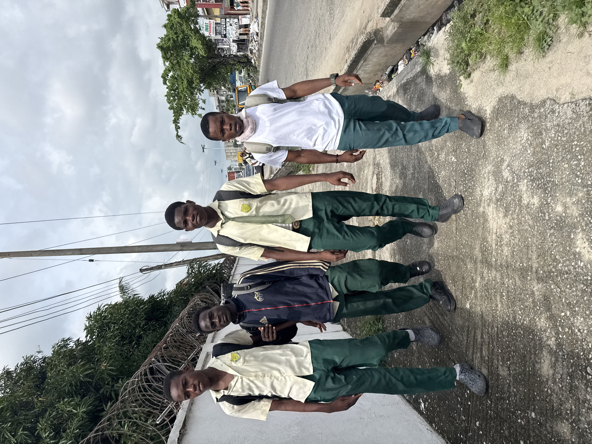 Four young men in school uniforms are walking along a sidewalk in Ado, Nigeria. The two young men on the left are wearing cream-colored shirts with green ties and green pants, while the two young men on the right are wearing cream-colored shirts with green ties and green pants, and one is wearing a white t-shirt with green pants and a scarf around his neck. They are all carrying backpacks. The sidewalk is concrete and appears somewhat weathered, with some debris and vegetation growing alongside it. In the background, there is a busy street with cars, motorcycles, and buildings. The sky is overcast. The environment suggests an urban setting in Nigeria.