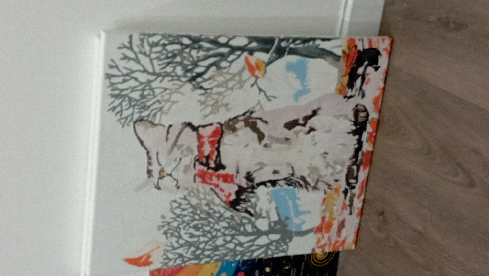 The image depicts a square canvas painting resting against a white wall adjacent to a wooden floor. The painting features a stylized image of a cat rendered in shades of grey, white, black, red and blue amidst abstract tree branches. There are hints of orange and yellow accents as well. The scene lacks sufficient details to place it within Nha Trang Ward, Vietnam.