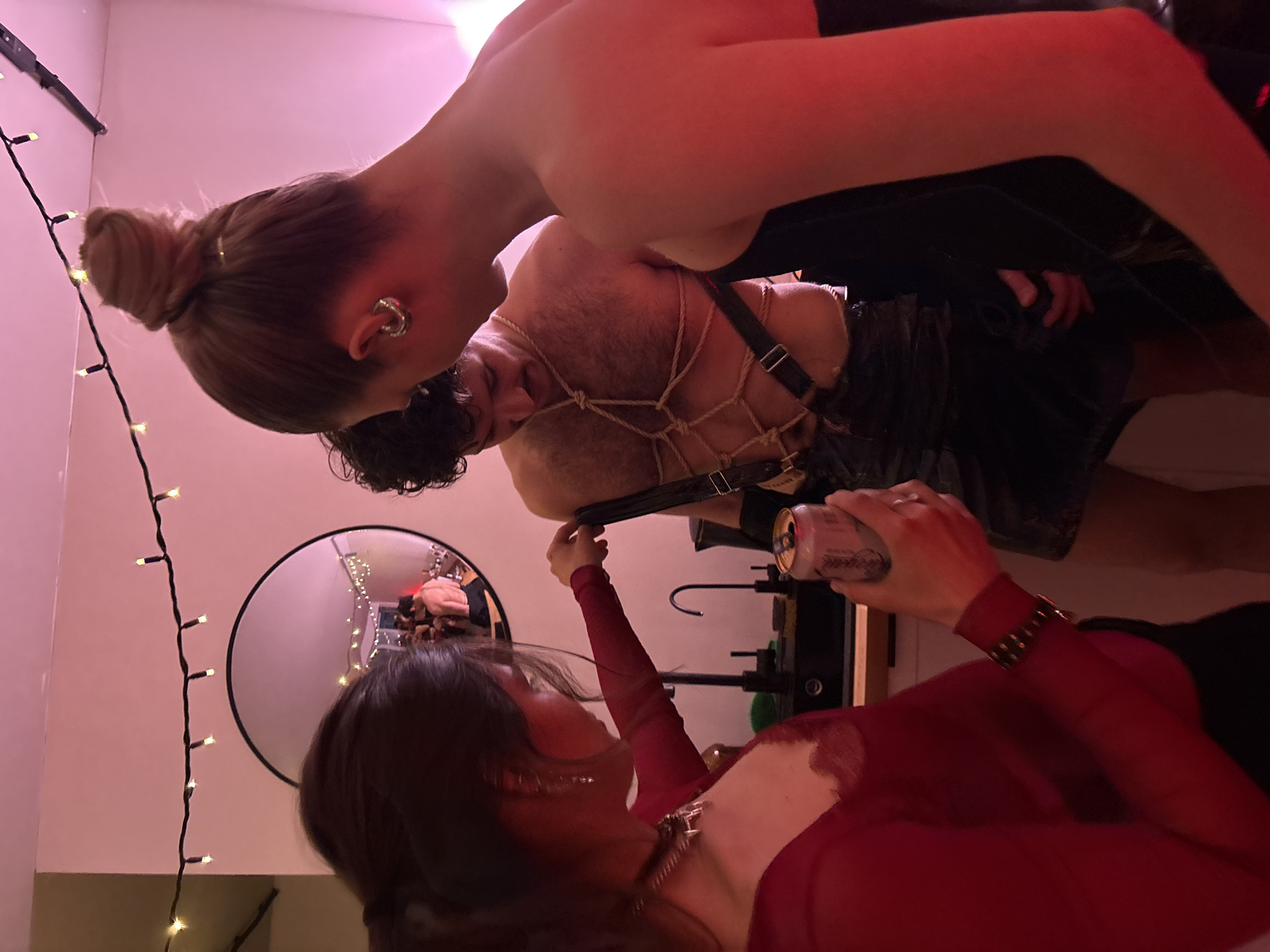 The photograph depicts an indoor scene with three individuals. One individual is suspended with ropes and harnesses. Another individual, wearing a red garment and a collar, holds a can. A third individual with their hair in a bun is positioned in the foreground. The setting includes a mirror on the wall, string lights, and what appears to be exercise equipment. The location is not discernible as Moscow, Russia, based on the visual elements provided.
