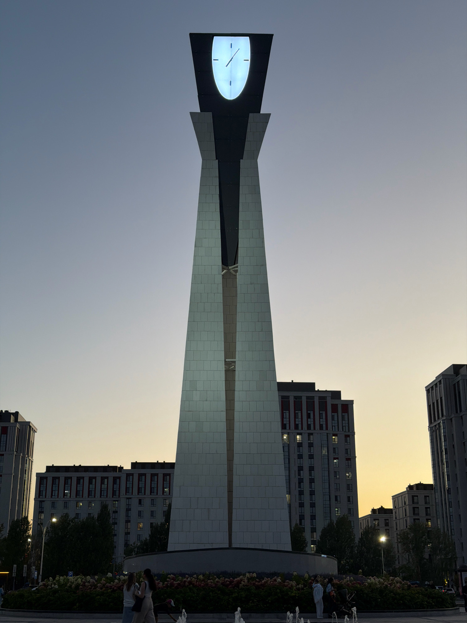 The image depicts the "Astana Clock Tower" located in the city of Astana, Kazakhstan, during twilight. The clock tower is a prominent, tall, modern structure with a large, illuminated clock face at its apex, displaying the time as approximately 3:03. The tower is primarily white with dark accents and a sharp, angular design. In the background, several modern residential buildings with red and gray facades are visible. The sky exhibits a gradient of twilight colors, transitioning from a clear blue at the top to a warm yellow-orange near the horizon. In the foreground, there is a flowerbed with a variety of colorful flowers and some people are walking in the area, including two women in the lower left corner of the frame and a figure in the lower right corner. Fountains are also visible in the lower part of the image.