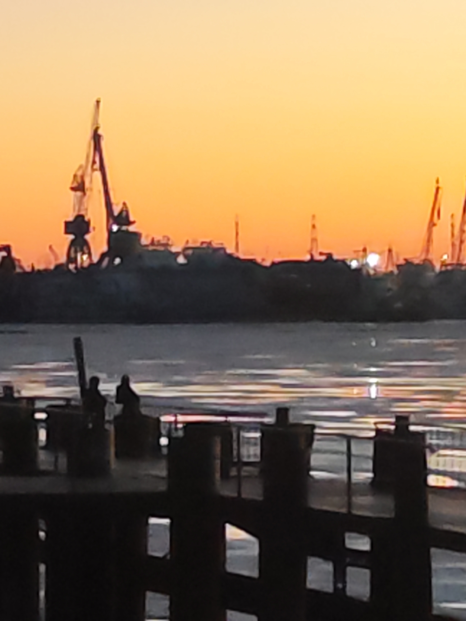 The image depicts a waterfront scene at dusk or dawn. In the foreground, there is a dark pier or dock with railings. Two silhouetted figures are visible on the pier. In the background, a body of water reflects the light from the sky. The skyline shows a cluster of industrial cranes and buildings, likely a port area. The sky is a gradient of yellow and orange. The location is within the city of Unknown, Unknown.