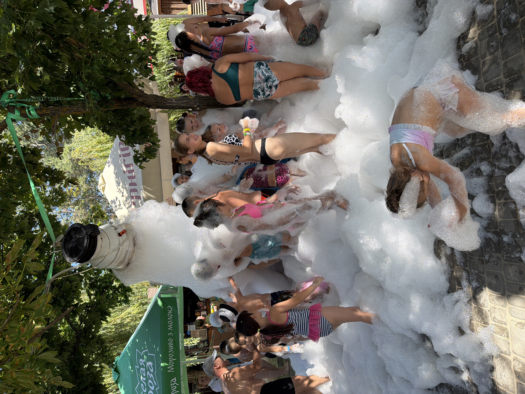 In Mar'yanivka, Ukraine, children and adults are gathered in a large pile of white foam, likely from a foam machine that is partially visible hanging from a tree branch. The event appears to be outdoors, with trees providing shade and tents set up in the background. People are dressed in swimwear, and some are interacting with the foam, playing and posing for the camera. The atmosphere seems festive and playful.