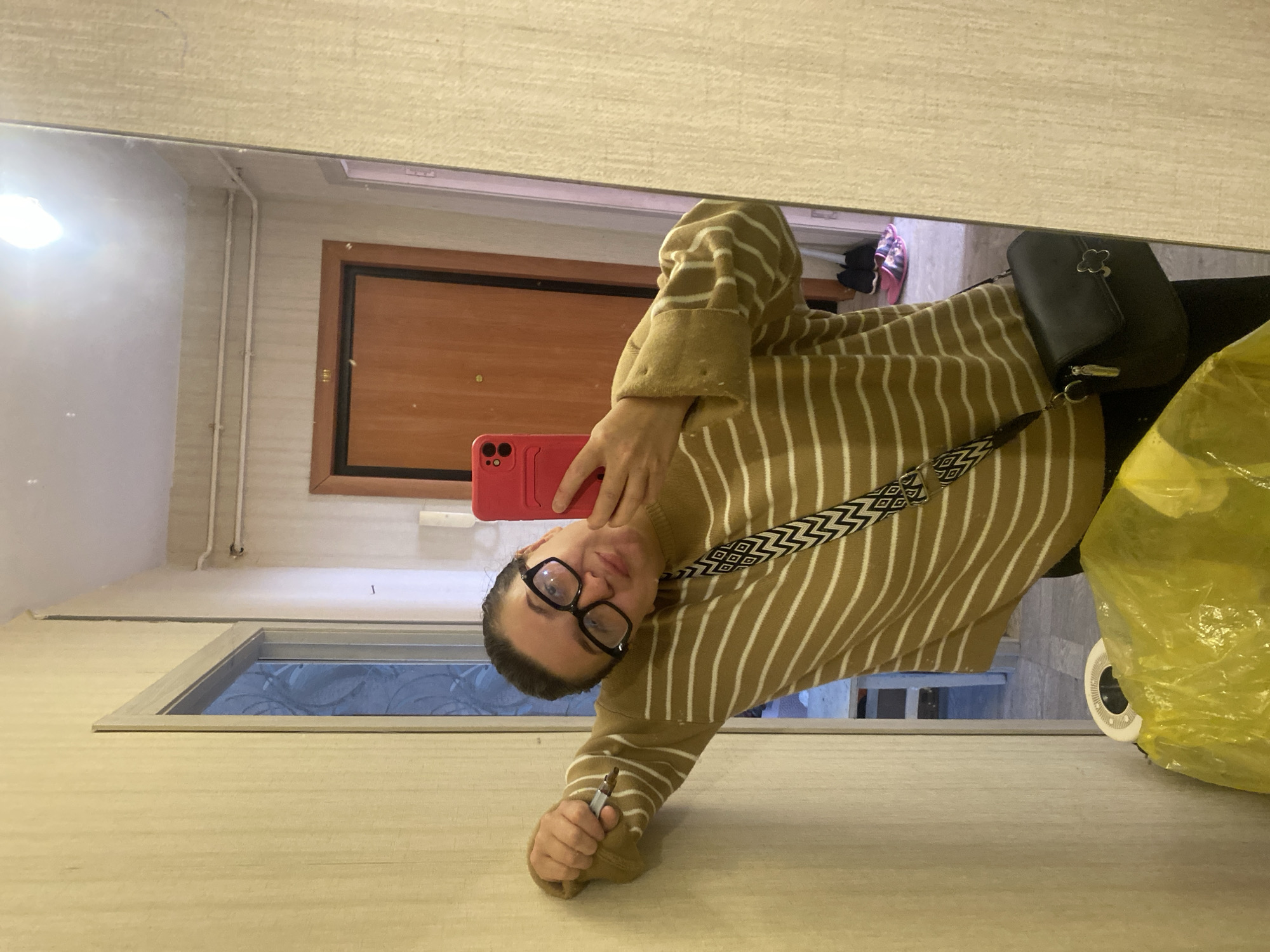 A young woman wearing a tan and white striped sweater, black-rimmed glasses, and a black crossbody bag takes a mirror selfie. She is leaning against a light yellow wall, holding a red smartphone in her right hand and a pen in her left. She is looking at the camera with a playful expression. In the background, a wooden door and a doorway leading to another room are visible, along with shoes on the floor. The image appears to be taken inside a residential hallway or room, with typical apartment building fixtures like baseboard heating and textured walls. There is no specific indication of the city of Chaikovsky, Russia, within the image itself.
