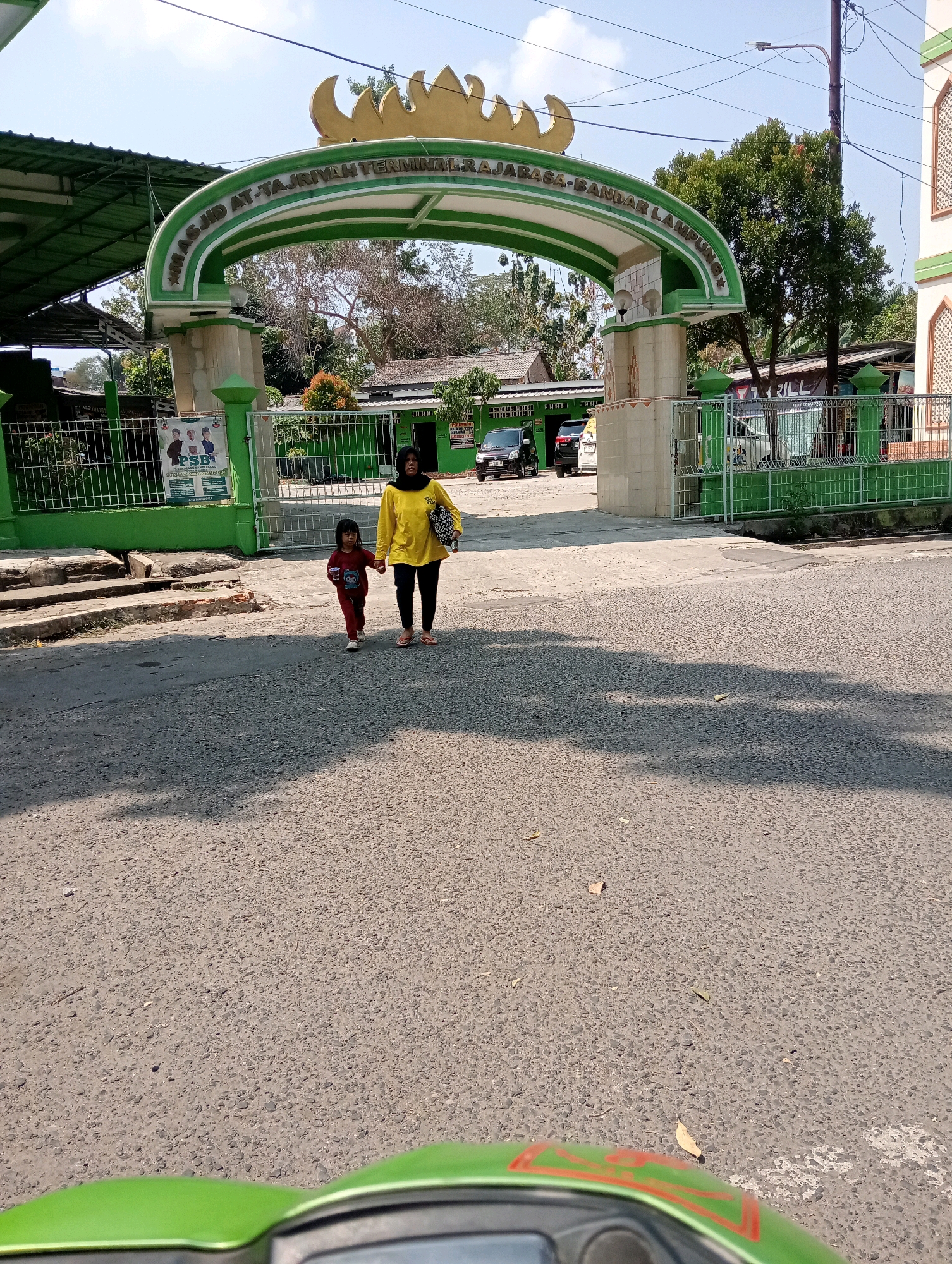 The image captures an outdoor scene in front of a mosque, identified by the Arabic inscription "MASJID AT-TAJRIYAH TERMINAL RAJABASA - BANDAR LAMPUNG" on an arched gateway. A woman wearing a yellow long-sleeved shirt, dark pants, and a black headscarf is walking hand-in-hand with a young child in a red shirt and pants. They are on a paved road, casting shadows from the bright sunlight. Behind them, a green fence encloses the mosque's grounds, where several cars are parked, including a black sedan and a white van. The environment appears to be a bright, sunny day.