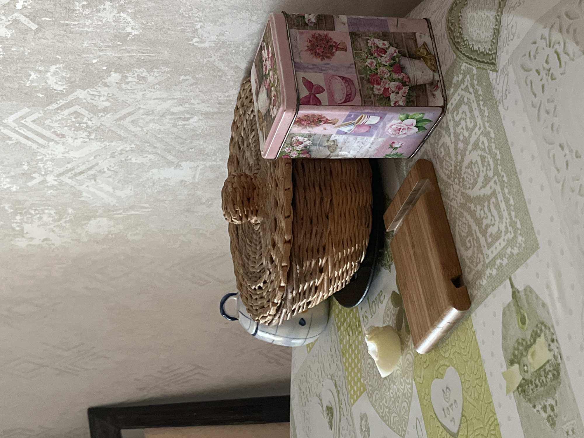 A still life photograph features a woven basket with a lid, a decorative tin, a ceramic jar, a wooden object, and a piece of food, all arranged on a patterned tablecloth. The background consists of a textured wall with a subtle geometric pattern. The scene appears to be indoors, possibly in a home setting. The lighting suggests it is daytime, with light casting shadows on the objects.