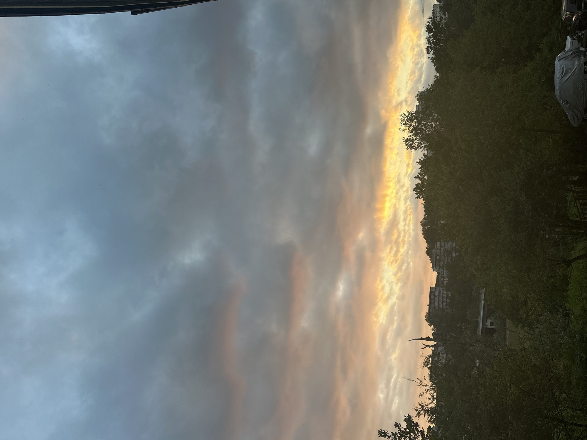 This media file captures a sunset scene in Razdolnoye, Russia. The image is oriented vertically, with the sky occupying the majority of the frame. Dramatic clouds are illuminated by the setting sun, creating a vibrant band of orange and yellow light on the right side, contrasted with darker, grey clouds on the left. Silhouetted trees and buildings line the horizon, suggesting an urban or suburban setting. The trees appear lush and green, indicating a season other than winter. A car covered with a tarp is visible on the far right. A few small birds are spotted in the sky. The overall mood is serene and picturesque, indicative of an evening twilight.