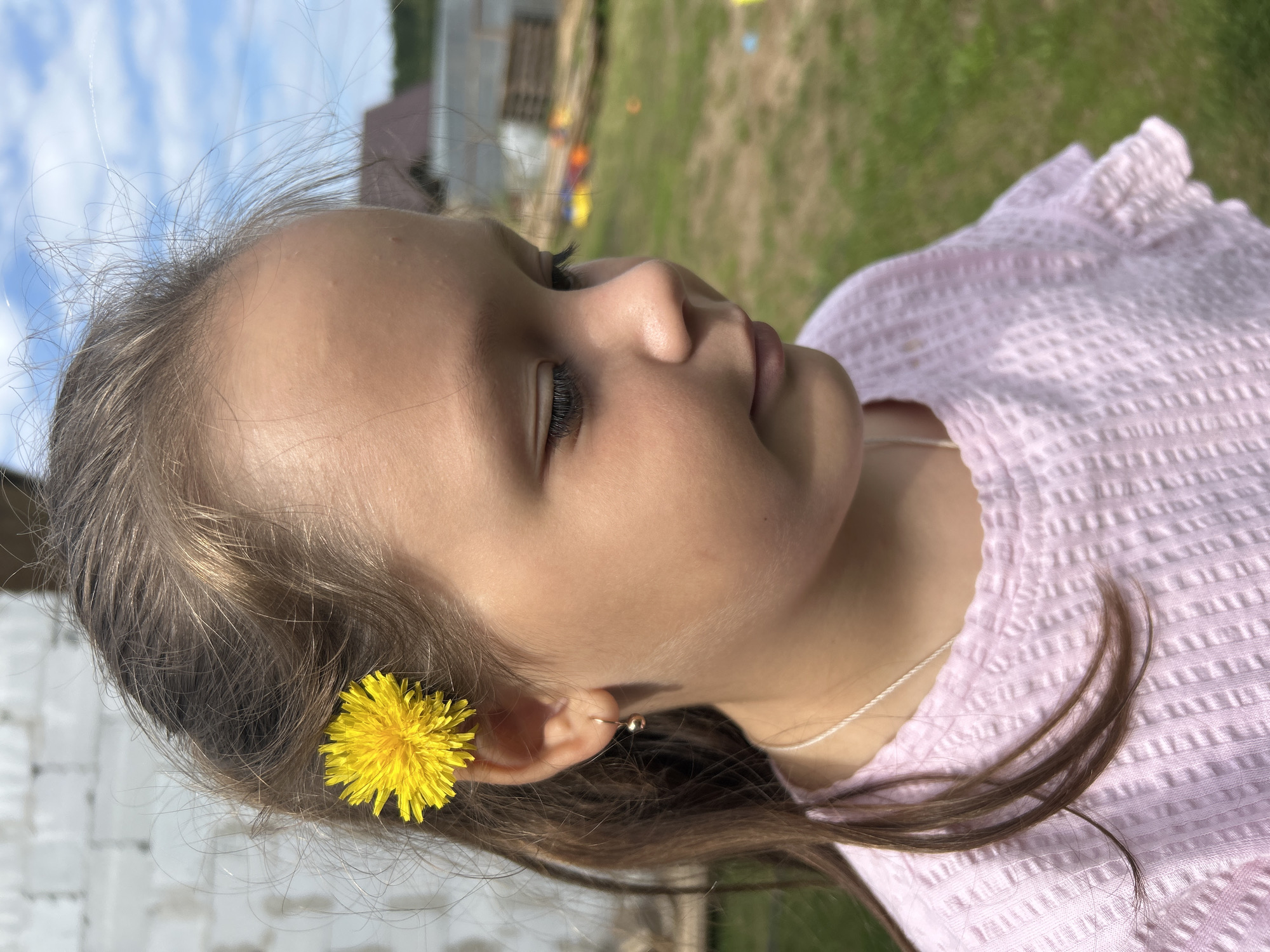 In a medium shot, a young girl with brown hair is positioned in the foreground, facing to the right. A bright yellow dandelion is tucked behind her right ear, and a delicate gold earring adorns her earlobe. She wears a pink, waffle-knit shirt with ruffled sleeves and a thin silver necklace. Her gaze is directed downwards, and her expression appears pensive. The background reveals a grassy area with some wooden structures and blurred colorful objects. The sky above is a clear blue with scattered white clouds. The image was taken outdoors, likely in a residential area or yard, possibly in Chaikovsky, Russia.