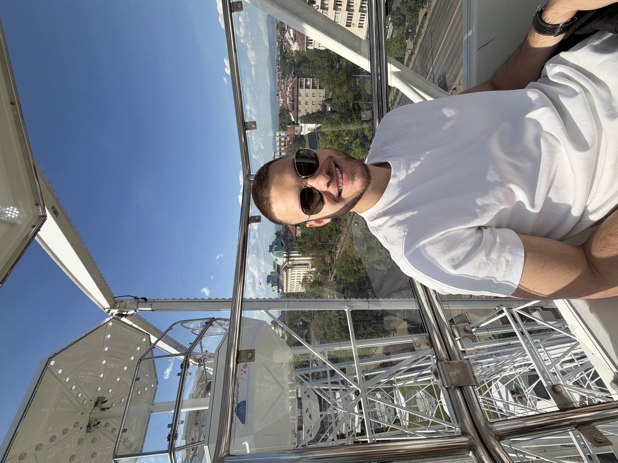 A man wearing a white t-shirt and sunglasses is inside a Ferris wheel cabin, smiling at the camera. Through the glass walls of the cabin, the cityscape of Sofia, Bulgaria, is visible.