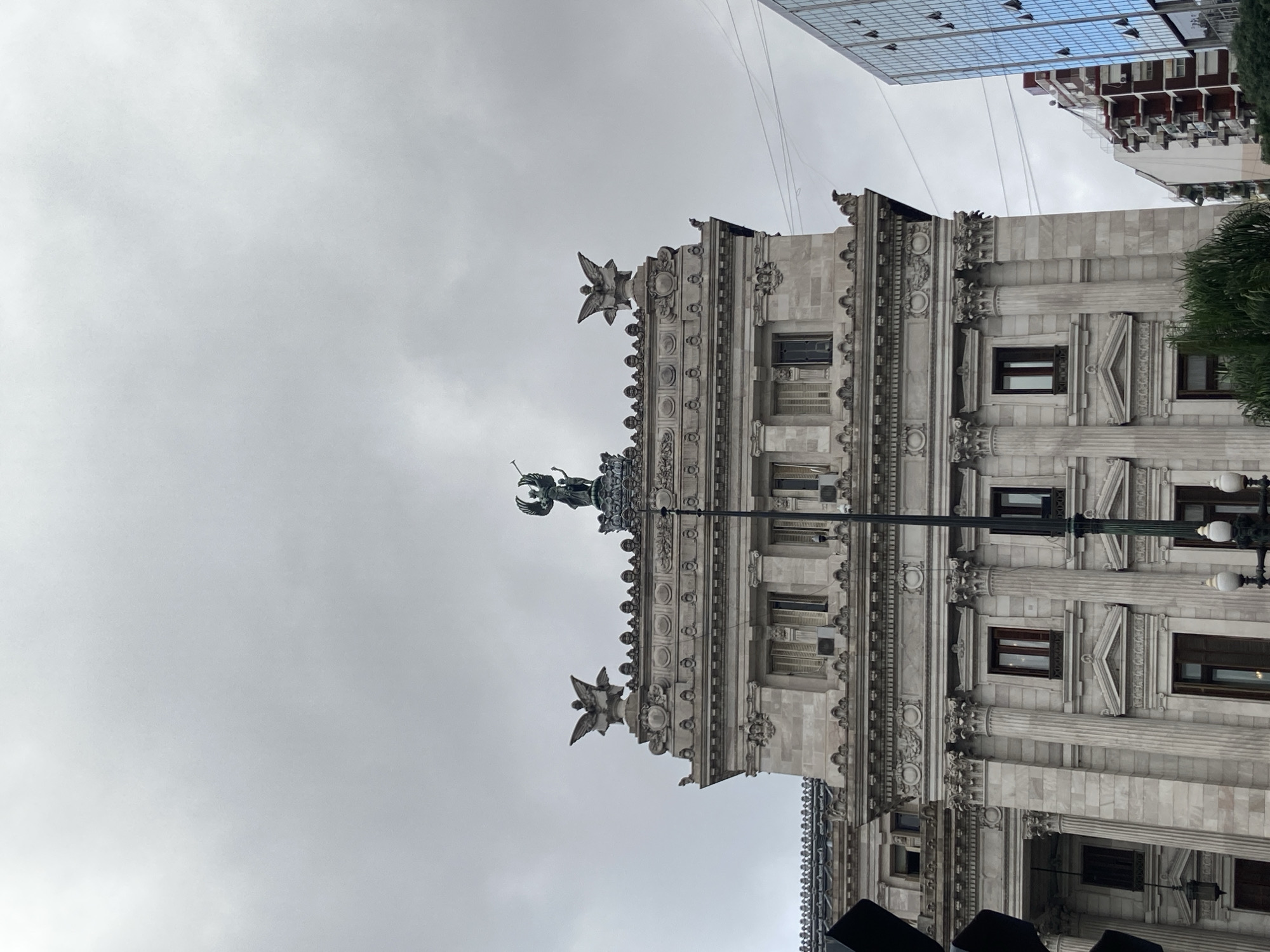 The image displays a grand, ornate stone building under an overcast, cloudy sky. The architecture is characterized by classical elements, featuring numerous fluted columns, elaborate cornices, and richly detailed friezes that adorn the facade. The building's light grey stonework is intricately carved, with decorative motifs around windows and along the roofline.
Prominently positioned on the roofline is a large central statue of a winged figure, possibly an angel or a classical goddess like Nike, holding an object while standing on a pedestal. Smaller, decorative winged figures, resembling eagles or mythical creatures, are also visible along the parapet. The building features multiple rectangular windows, some with dark frames.
In the upper background, a modern high-rise building with a blue-tinted glass facade reflects the cloudy sky, creating a contrast with the historical structure. Another building with several small balconies is partially visible behind. A tall, dark street light pole with multiple lamps stands in the mid-ground, slightly obscuring parts of the main building. A patch of green foliage, likely from a tree or bush, is visible on the right side of the frame. The scene suggests an urban environment where historical grandeur meets contemporary architecture. It is daytime, but the weather is cloudy, resulting in soft, diffused light. There is no discernible activity or visible text in the image.