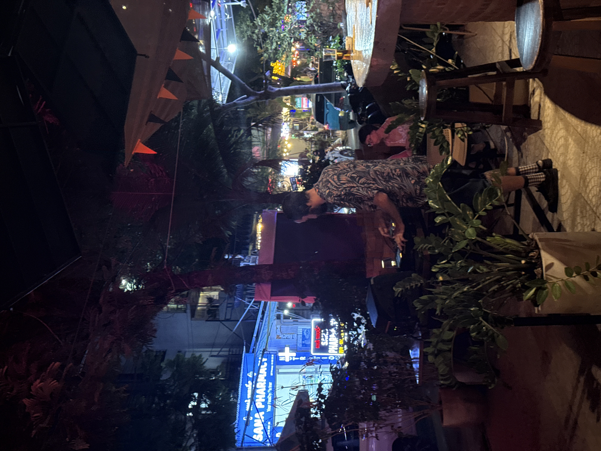 The image depicts a night scene at an outdoor establishment in Đà Nẵng, Vietnam, likely a bar or restaurant. A man with a beard, wearing a short-sleeved shirt with a tropical print, is seen standing at a DJ booth, appearing to be operating equipment. His hands are visible near a mixing board. In the background, a blue sign with "SANA PHARMACY" is illuminated, indicating a nearby pharmacy. Further in the distance, the street is visible with illuminated signs and vehicles, including a blue car. A woman with glasses is seated at a table to the right, partially obscured by plants. Potted plants are strategically placed around the area. The overall ambiance is casual and festive, with string lights and decorations suggesting a social gathering. The flooring appears to be tiled.