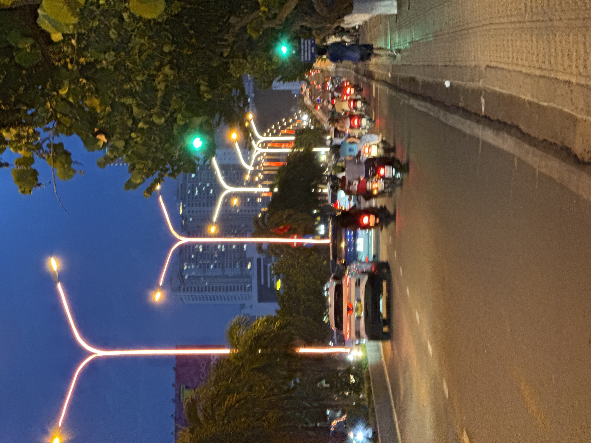 The image depicts a street scene in Nha Trang, Vietnam, at dusk or night. A multi-lane road is visible with traffic, predominantly motorbikes, moving in both directions. Streetlights with bright orange-red illumination line the road, casting a warm glow on the asphalt. Tall buildings with many windows are visible in the background, suggesting an urban environment. Lush green trees with large leaves flank the right side of the road, and some palm trees are present on the left. A sidewalk runs along the right side, where a few individuals are walking. The traffic lights are green in the visible sections. The overall impression is a busy city street at night, with a mix of vehicles and pedestrians.