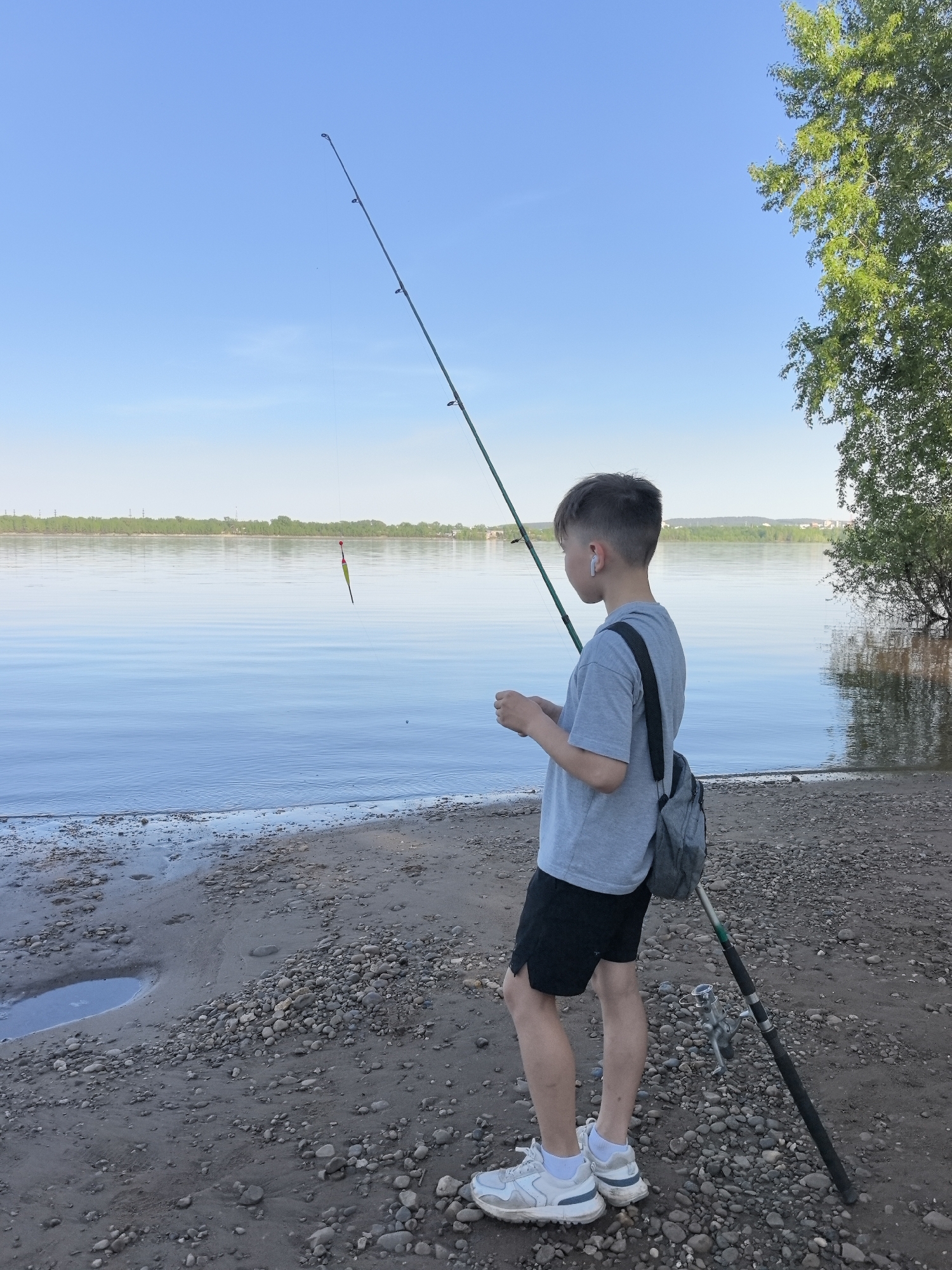 A boy stands on a gravelly beach in Noviy, Russia, holding a fishing rod with a bobber in the water. He wears a light grey t-shirt, black shorts, white sneakers, and a grey bag over his shoulder, with a white wireless earbud in his ear. The fishing rod leans against his leg, with the reel visible. In the background is a wide river, with trees lining the opposite shore. To the right, a large tree grows near the water's edge. The sky is blue and clear.
