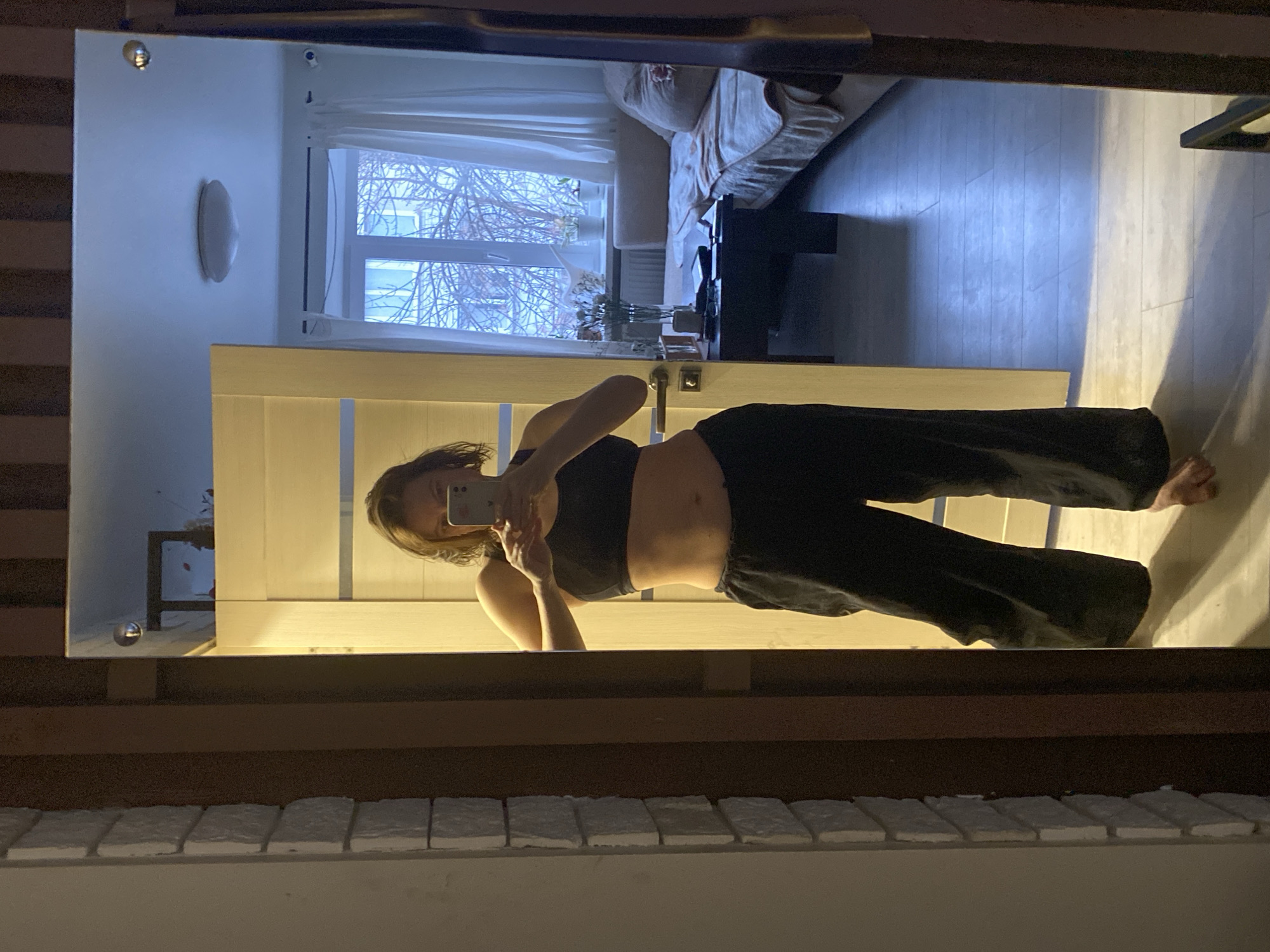 A mirror reflects a woman taking a photo of herself. She is wearing a black sports bra and loose black pants. The reflection shows a portion of a room with a couch, a window, and what appears to be a radiator or vertical decorative panel. Through the window, bare tree branches can be seen. The exterior structure visible at the bottom of the image has a row of white rectangular shapes on top, which suggest some sort of construction (like bricks or stone), then a dark brown band and the mirror on top. The setting cannot be confirmed as Chaikovsky, Russia based on the image. There's no information that confirms location.