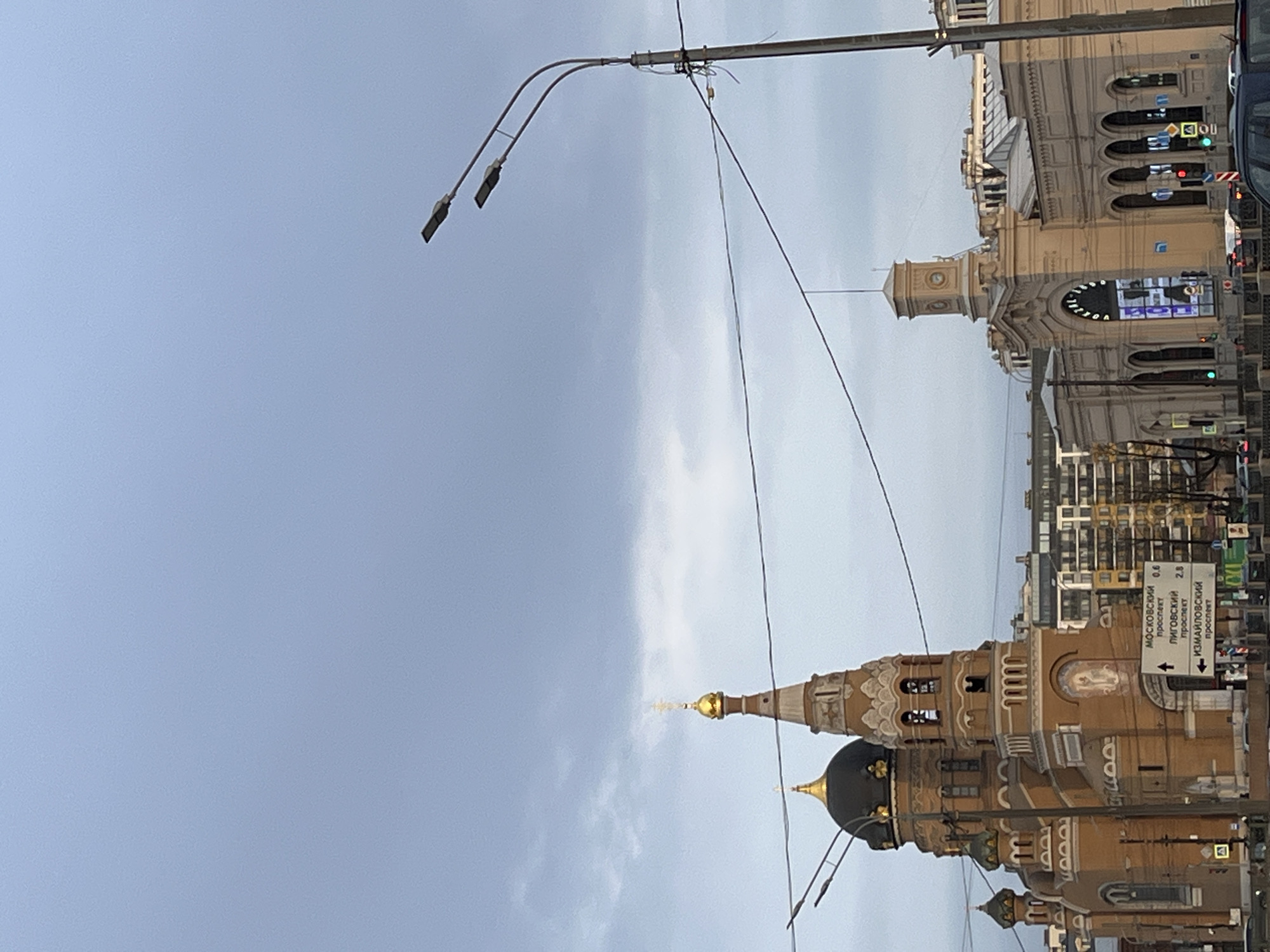 The image depicts a street scene in a city of unknown location. The sky is overcast with a range of light to darker gray clouds. A street lamp is present in the upper left corner. In the foreground, there is a large architectural structure with a black dome and gold spire. The building’s facade features arched windows and decorative detailing. Behind this building is a street sign, displaying text in Cyrillic alphabet. Further down the street, there is another building with a clock tower and detailed architecture. Traffic lights and a partial view of a vehicle are also visible.