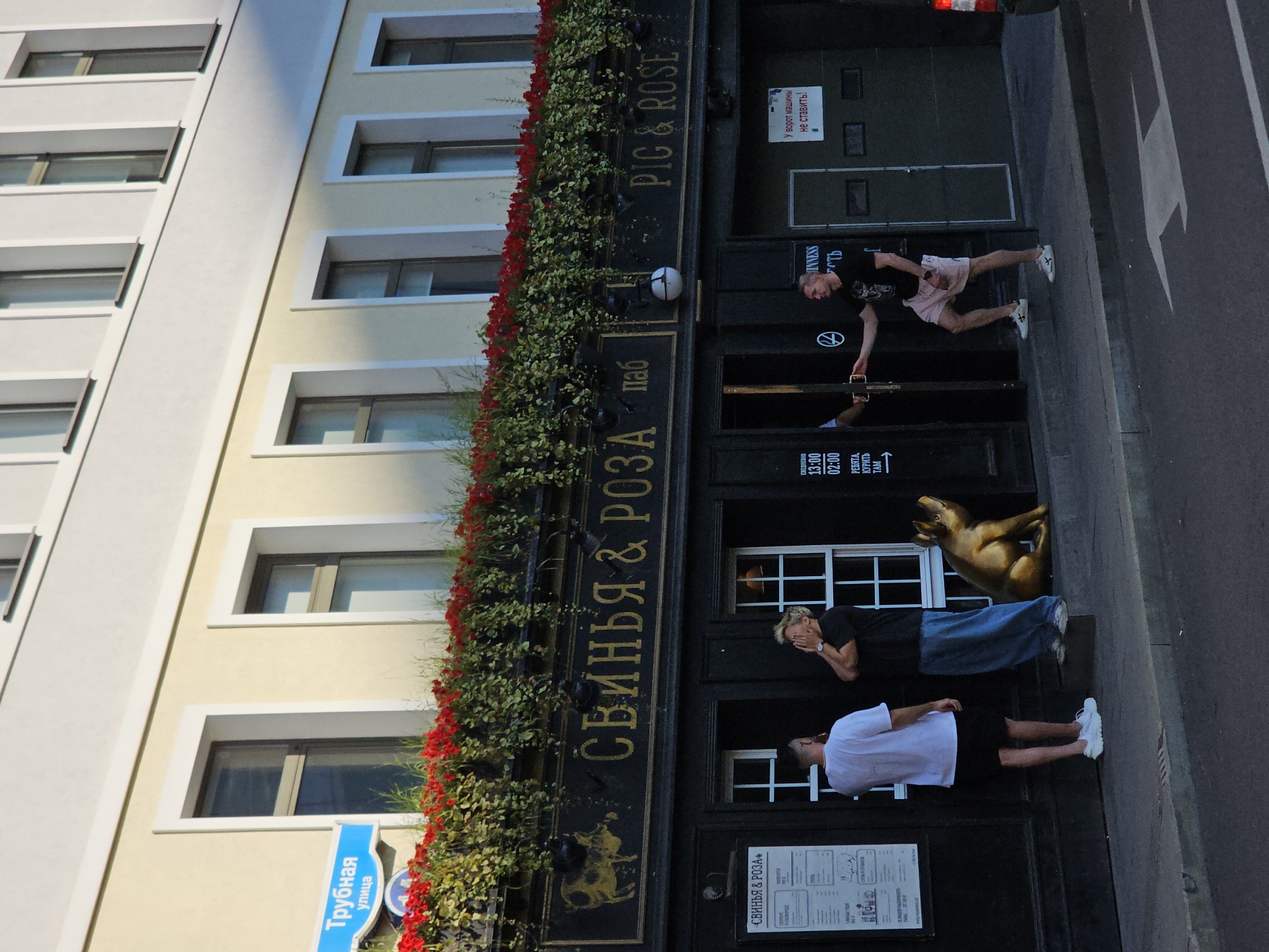 The image captures the exterior of a pub named "Pig & Rose" in Moscow, Russia, specifically on Trubnaya Street. The pub's facade is dark, with gold lettering displaying its name in both Russian ("СВИНЬЯ & РОЗА") and English ("PIG & ROSE"). Above the pub's entrance, an awning is adorned with lush greenery and vibrant red flowers.
Several people are present on the sidewalk in front of the pub. On the left, two individuals stand near a menu board. One, wearing a white t-shirt and black shorts, faces away from the camera, while the other, in a black t-shirt and blue jeans, covers their face with their hands. To the right of these individuals, a golden statue of a pig is seated. Further to the right, a man in a black t-shirt and pink shorts is interacting with the pub's entrance, appearing to hold the door open for someone inside. A sign with Russian text, "У ворот машины не ставить!", which translates to "Do not park cars at the gate!", is visible on the wall to the right of the entrance. The street in the foreground shows a white directional arrow painted on the asphalt. The building itself is light-colored with multiple windows.