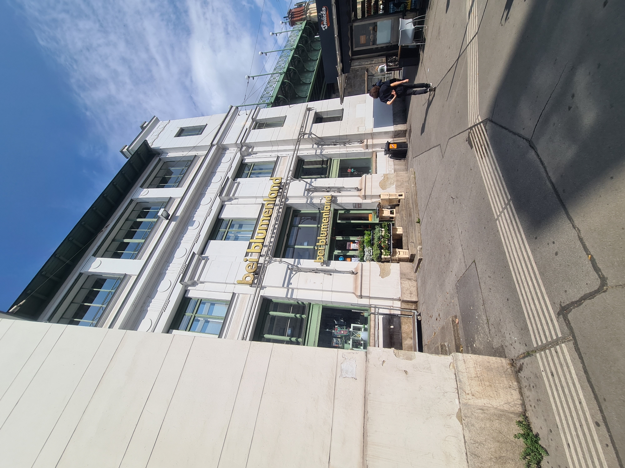 The image shows the exterior of a building in Vienna, Austria, with the signage "beiblumenland" visible. The building is white with green-trimmed windows and appears to be a commercial establishment. In the foreground, a person is walking away from the camera, wearing a black shirt and dark pants. To the right of the building, there is a green bridge structure and a "Snacks" kiosk. The scene is set on a sidewalk with tactile paving. The sky is blue with scattered clouds, and the sun is casting shadows.