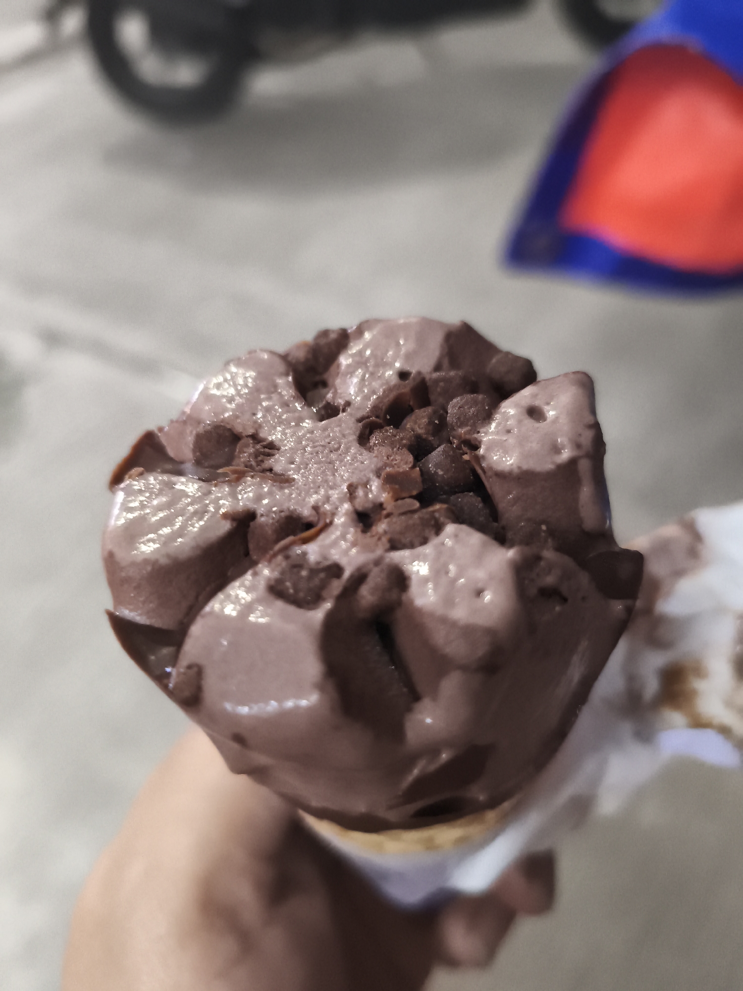A close-up shot shows a chocolate ice cream cone covered in chocolate chips being held in a person's hand. The ice cream appears slightly melted and glossy. The background is blurred, but a motorcycle and what appears to be a blue and red object are discernible. The image was likely taken in Depok, Indonesia.