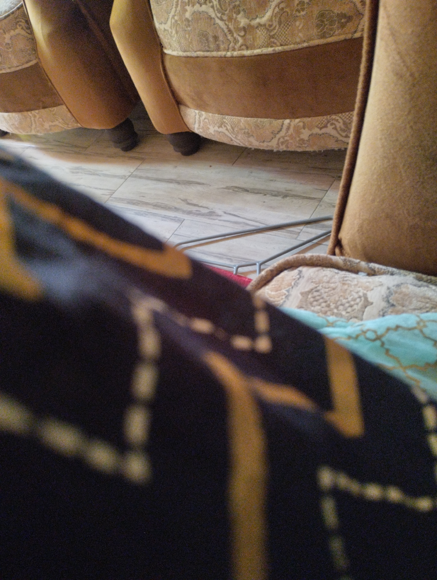 The image shows the lower section of two chairs or a sofa. The chairs have brown upholstery with a patterned beige fabric trim. The flooring underneath appears to be light-colored tile with a subtle wood grain pattern. In the foreground, a dark fabric with a yellow geometric pattern is draped over something, partially obscuring the view of the chairs. A thin metal rod is visible across the floor between the chairs. The scene is indoors, with no indication of its specific location in the city.