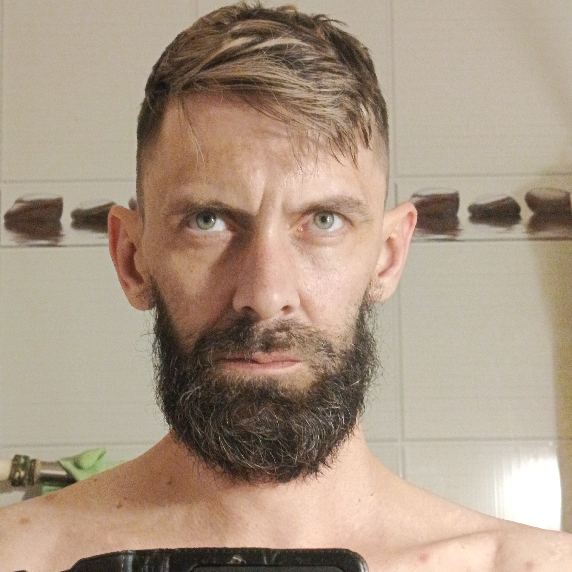 A man with light brown hair, a beard, and green eyes is centered in the frame. He is shirtless. The background is a bathroom, featuring white tiles with a horizontal row of brown decorative tiles near the top. A towel rack with a green item is visible in the lower-left corner. Part of a black object is in the foreground. The location is Марковский, Russia, though the image does not provide any details to confirm it.