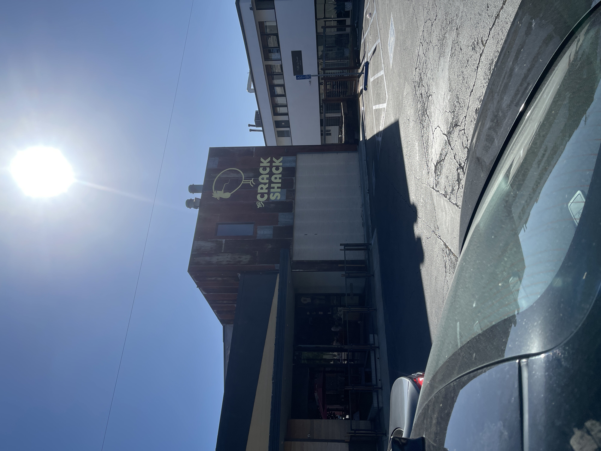 The image shows the building of "Crack Shack", a restaurant known for its chicken dishes, in Costa Mesa, California. The building has a weathered, rusty appearance and the restaurant's logo prominently displayed on it. A car is partially visible in the foreground, with the restaurant situated on the left side of the frame. The parking lot and an adjacent building are also in view. The bright sunlight illuminates the scene.