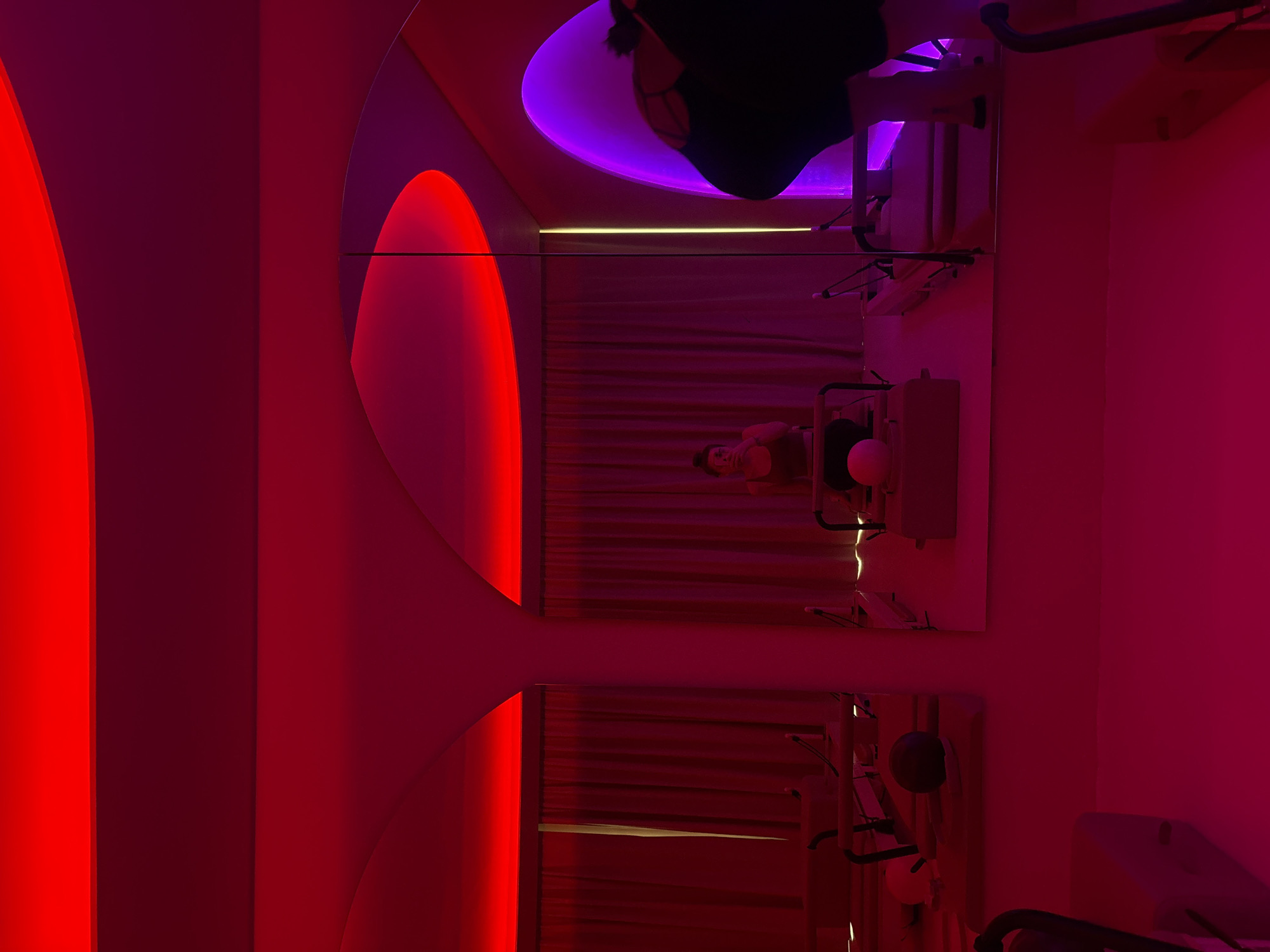 The image depicts an interior space in Mexico City, Mexico, illuminated with a mix of red, purple, and white light. Two vertically stacked mirrors on the wall reflect a curtained area where a person is sitting. Medical or cosmetic equipment is visible, suggesting a clinic or treatment room. Additional equipment and objects hang on the walls around the mirrors.