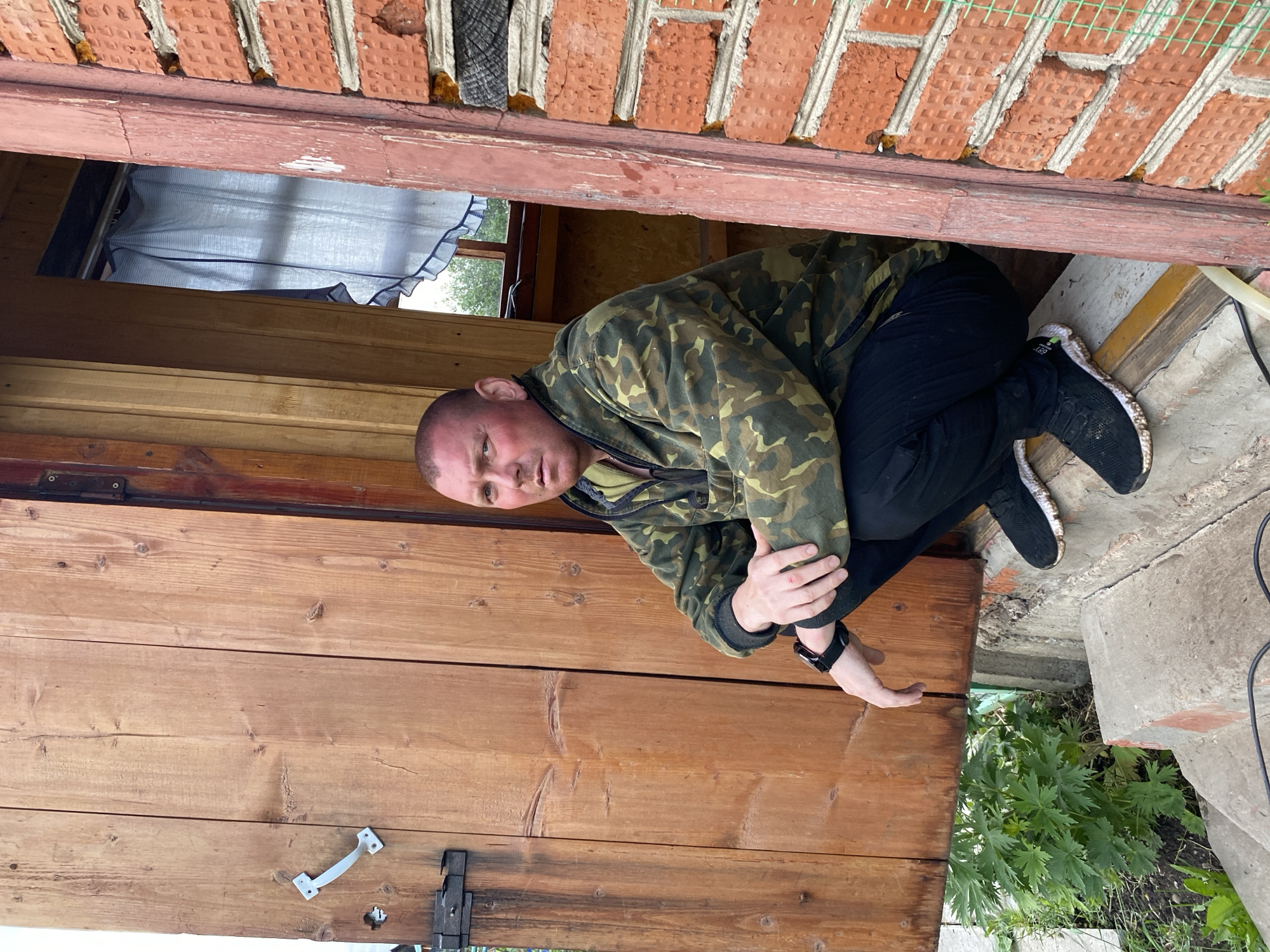 An adult male is observed squatting in an open doorway in Chaikovsky, Russia. He is positioned on a concrete step or threshold, facing forward towards the camera. The man has a shaved head, fair skin with a sun-reddened complexion on his face and ears, and a neutral facial expression. He wears a camouflage-patterned hooded jacket or pullover in green, brown, and black tones, dark-colored pants, and black athletic shoes with white soles. A black digital watch is visible on his left wrist.
To his left is a wooden door or wall section constructed from vertical planks, featuring a white, curved pull handle and a dark metal latch assembly. Directly behind the man, through the doorway, a window with a light-colored, ruffled curtain (blue and white striped) is visible, with green foliage apparent outside. To his right, a red brick wall with visible mortar lines and minor damage/repairs (orange sealant, green mesh) forms part of the structure. Concrete steps descend from the doorway. To the left of the steps, green vegetation, including a large-leafed plant, grows in the soil. A dark electrical cord is visible on the lower steps.