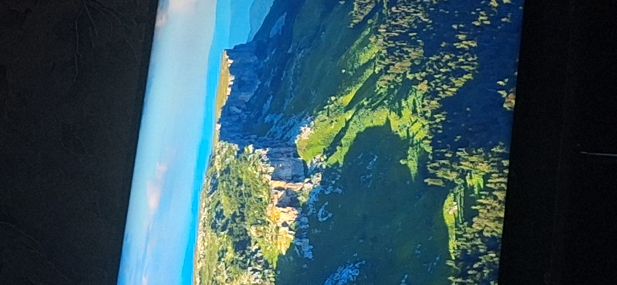 The image displays a vast, mountainous landscape under a bright sky with scattered clouds. The terrain features steep, rocky cliffs with a flat summit, covered in lush green vegetation and trees. The sunlight casts dramatic shadows across the slopes, highlighting the rugged topography. In the lower portion of the image, a dense forest stretches across a hillside, with a small clearing containing a few structures that appear to be houses or cabins. The overall scene conveys a sense of natural grandeur and remoteness. The location is not in the city of Bila Tserkva, Ukraine, as this is a natural landscape.