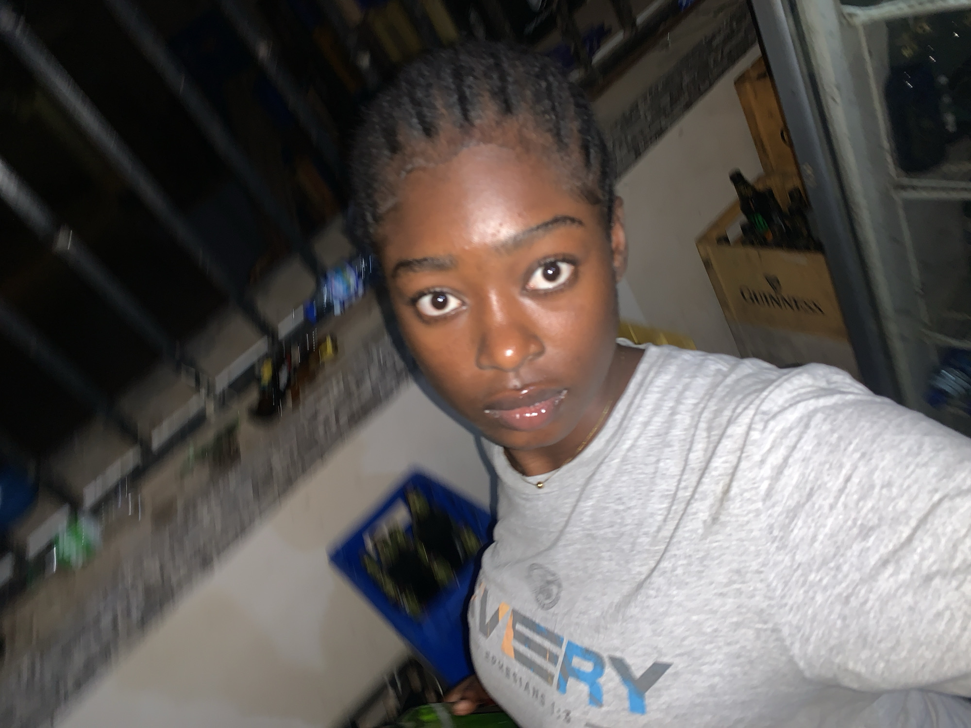 A young woman with dark skin and braided hair is pictured in a close-up, slightly tilted selfie. She is wearing a light grey t-shirt with text on it.
Behind her is a stairwell, with metal bars visible in the background. Adjacent to the stairwell, on the wall, is a Guinness beer crate containing bottles. To the left, slightly below the woman, there is a blue object.
Based on available information, the picture may have been taken indoors, possibly inside the woman's home. There is insufficient visual information to confirm the exact location in Kabong, Nigeria. The environment suggests an urban residential area.