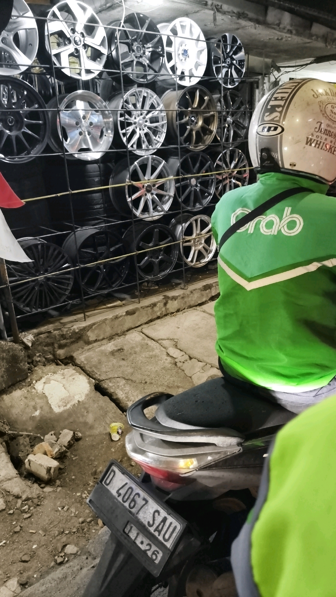 The image depicts an outdoor scene in a gritty urban environment, likely a roadside stall or workshop specializing in car rims and tires. In the foreground, a motorcycle is partially visible, featuring a license plate with the inscription "D 4067 SAU" and "11.26". A person wearing a bright green jacket with a "Grab" logo on the back and a helmet adorned with vintage whiskey branding is seated on the motorcycle, facing away from the camera. The background is dominated by a large display of car rims of various designs and colors, neatly arranged on shelves. The ground appears to be concrete, with some debris and broken bricks scattered around, suggesting a less than pristine setting. The overall atmosphere is utilitarian and functional, indicative of a business operating in a busy South Jakarta, Indonesia locale.