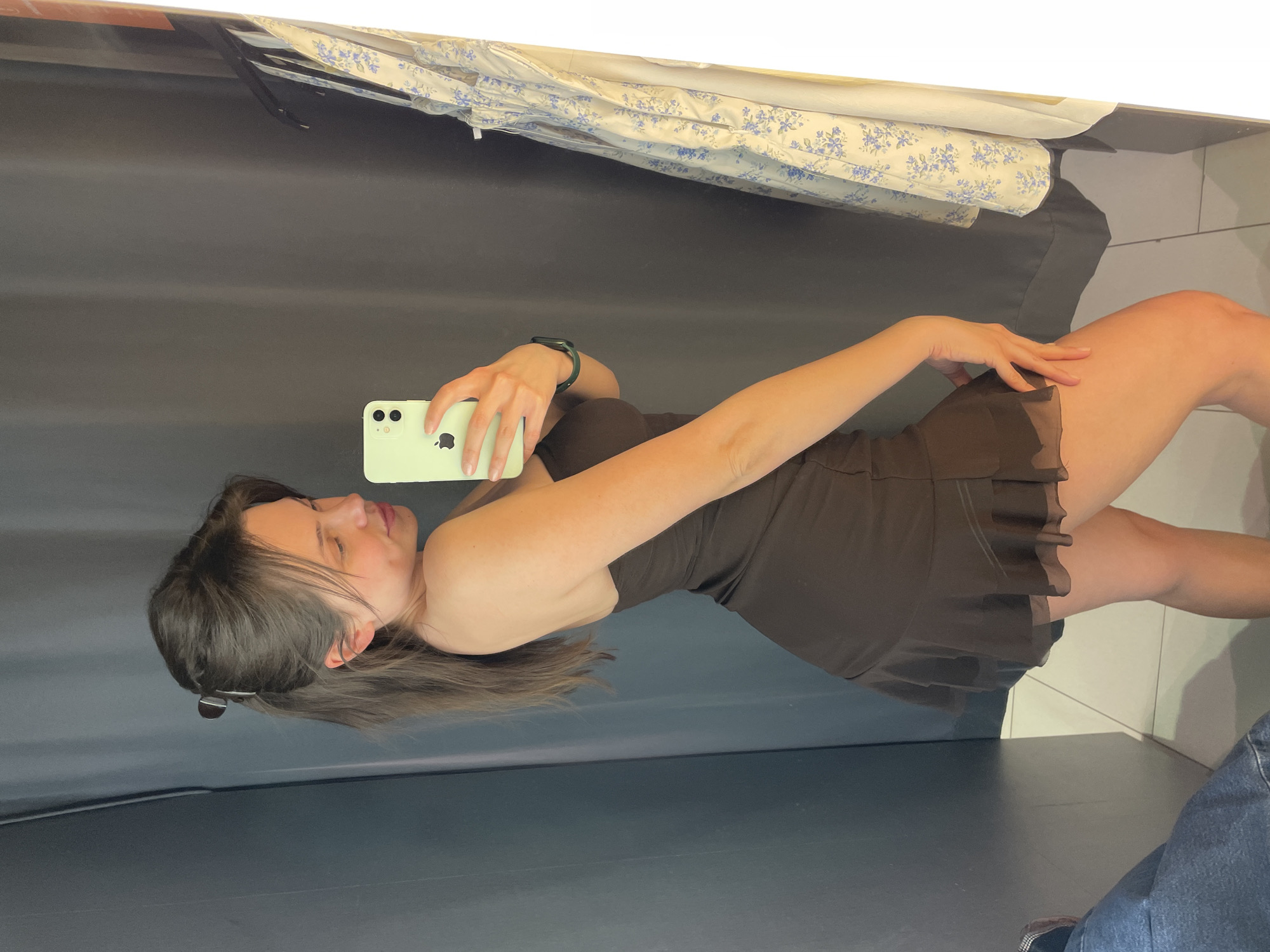 A young woman is depicted taking a mirror selfie with a light green iPhone, held in her right hand, showcasing the Apple logo on its back. The image is rotated 90 degrees counter-clockwise, presenting her in a horizontal orientation. She is wearing a dark brown or black strappy mini-dress with a subtly ruffled hem. A dark green smartwatch is visible on her left wrist. Her dark hair is pulled back with a hair clip, and she has a gentle expression, with her eyes slightly downcast or partially closed. Her left hand rests on her left thigh.
The background of the reflection reveals a dark, plain wall or curtain. Above this, a white surface, possibly a shelf or counter, reflects folded or rolled fabric adorned with a blue floral pattern against a white background. In the lower right corner, a partial view of another person's leg in blue jeans and a shoe is visible, suggesting either another individual present or a complex reflection within the space. The floor is dark and somewhat reflective. The setting appears to be an indoor space, likely a dressing room or a retail fitting area, located in Budapest, Hungary. The lighting is even and soft.