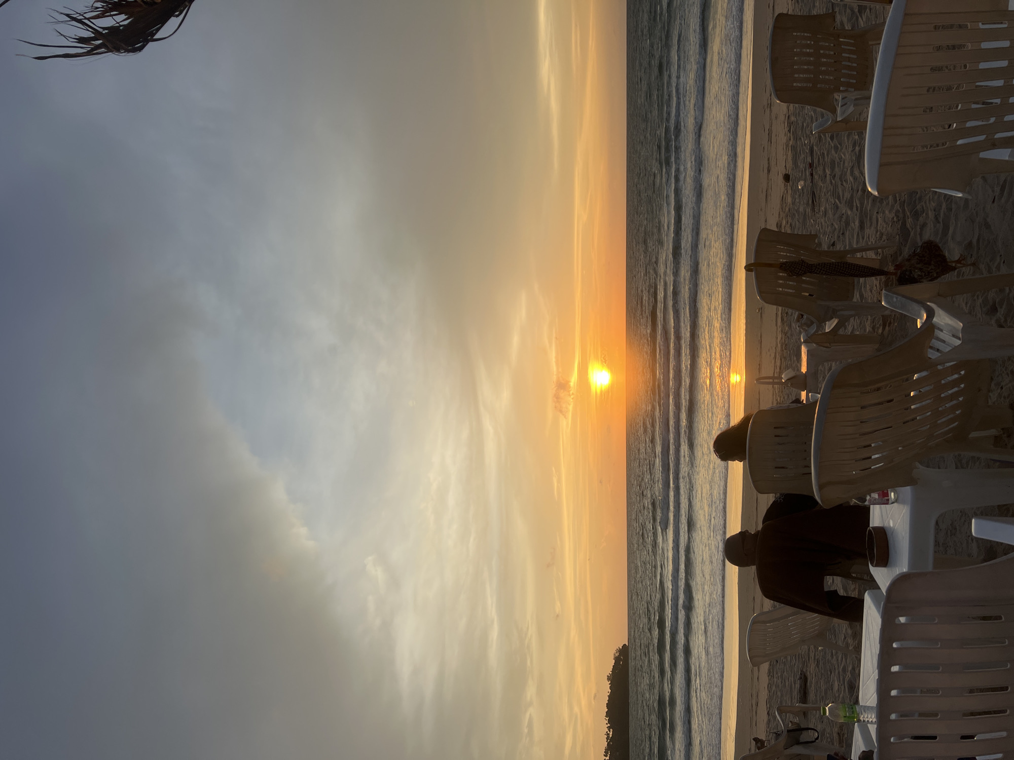 The image depicts a sunset scene on a beach in Choeng Thale, Thailand. In the foreground, several white plastic chairs and tables are set up on the sand, suggesting an outdoor dining or relaxation area. A few people are seated at one of the tables, facing away from the camera and towards the ocean. One person is wearing a dark brown garment over their shoulders. There are bottles of water and a small bowl on the tables.
The middle ground shows a sandy beach leading to the ocean. Waves are gently rolling onto the shore. In the distance, a tree-covered island or peninsula is visible on the left side of the frame.
The background is dominated by the sky and the sunset. The sun is a bright orange orb, positioned just above the horizon, casting a warm glow across the scene. The sky is filled with soft clouds that are tinged with orange and yellow hues. A frond of a palm tree is visible in the upper right corner, adding a tropical element. The overall atmosphere is serene and peaceful, characteristic of a sunset on a Thai beach.