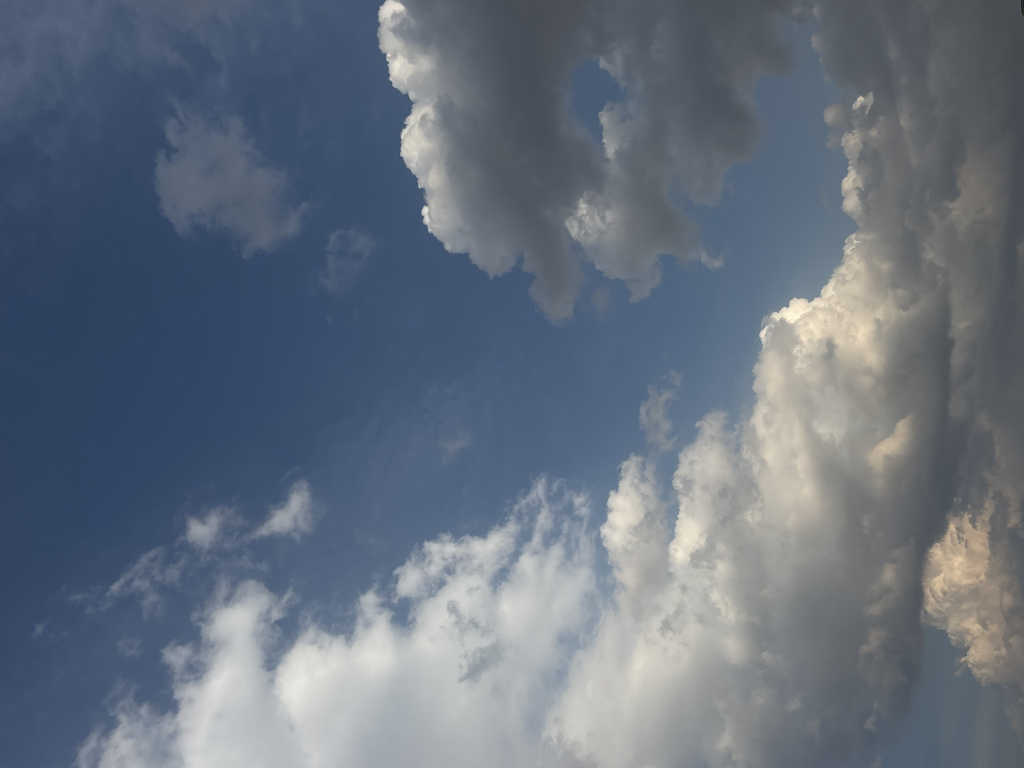 The image depicts a daytime sky with varying cloud formations. The left side of the frame features a clear blue sky with scattered wispy clouds. In the upper center and right of the image, denser cumulonimbus clouds are present, exhibiting shades of gray and white. Sunlight appears to be illuminating the edges of the clouds on the right, creating highlights. The scene captures an open sky with no visible ground elements or urban infrastructure. Although requested, there is no verifiable evidence in the image to determine the specific location in Puebla, Mexico. The image contains no people, identifiable objects, or actions beyond the natural appearance of the sky and cloud formations.