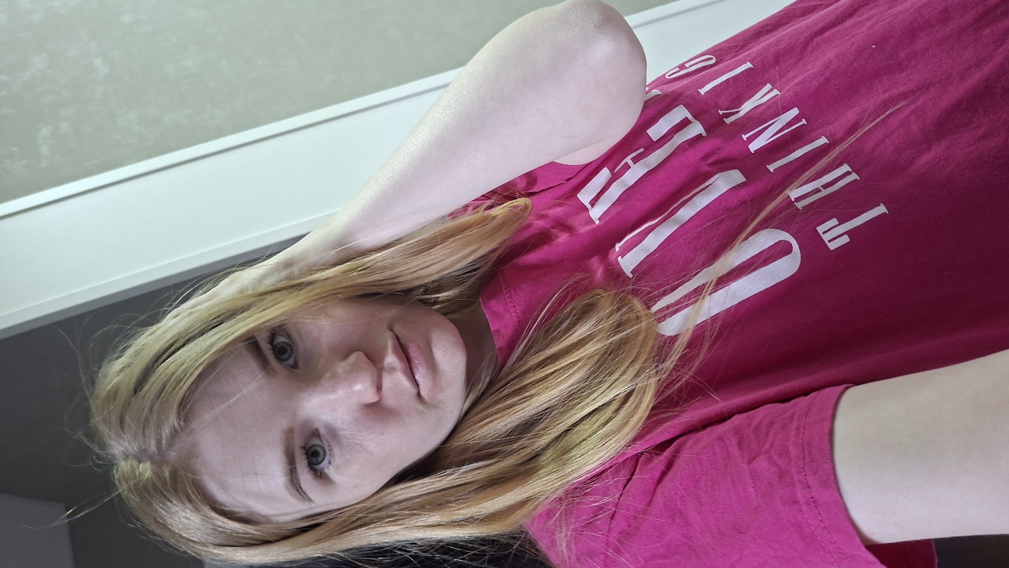 A blonde woman is depicted in an indoor setting. The frame captures her face and a portion of her upper body. She is wearing a bright pink T-shirt with white block letters that spell out the phrase "STYLE THINK." Her left arm is raised and positioned near her head. The background includes a white wall and a gray area, possibly part of the ceiling. The location in the city of Chaikovsky, Russia is not discernible from the image.