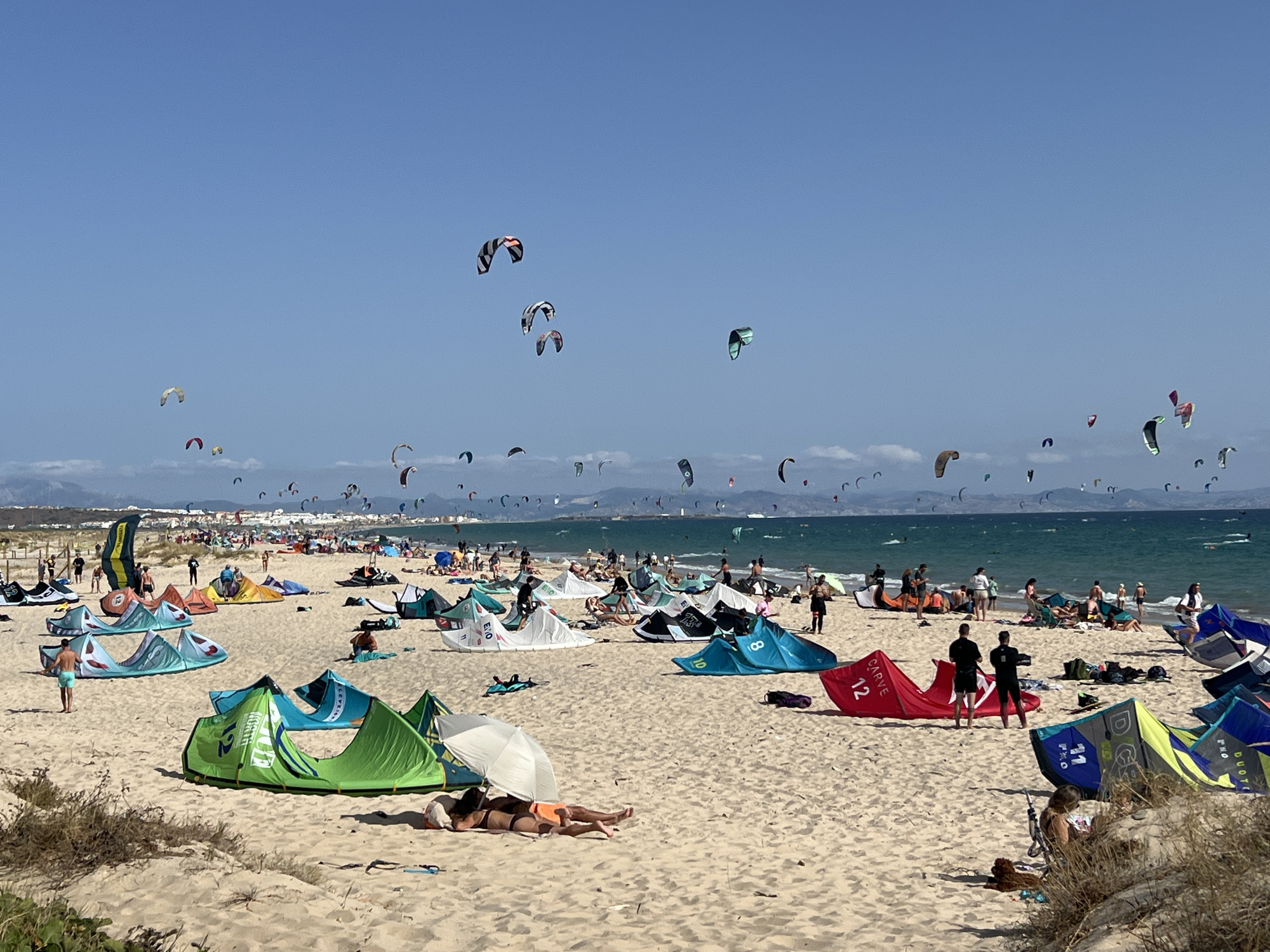 The image depicts a crowded beach scene in Tarifa, Spain, a location known for kitesurfing. The foreground is dominated by sand dunes with sparse vegetation and several colorful kitesurfing kites laid out on the sand, some set up as tents. Numerous people are visible on the beach, some sunbathing, others standing or walking. In the middle ground, more kitesurfing kites are set up, and a dense crowd of people are gathered. Beyond the beach, the turquoise sea is visible, filled with many kitesurfers in the water, their kites creating a vibrant spectacle against the clear blue sky. The distant background shows a coastline with buildings and hills. The overall impression is one of a lively and active beach day, with kitesurfing being the primary activity.