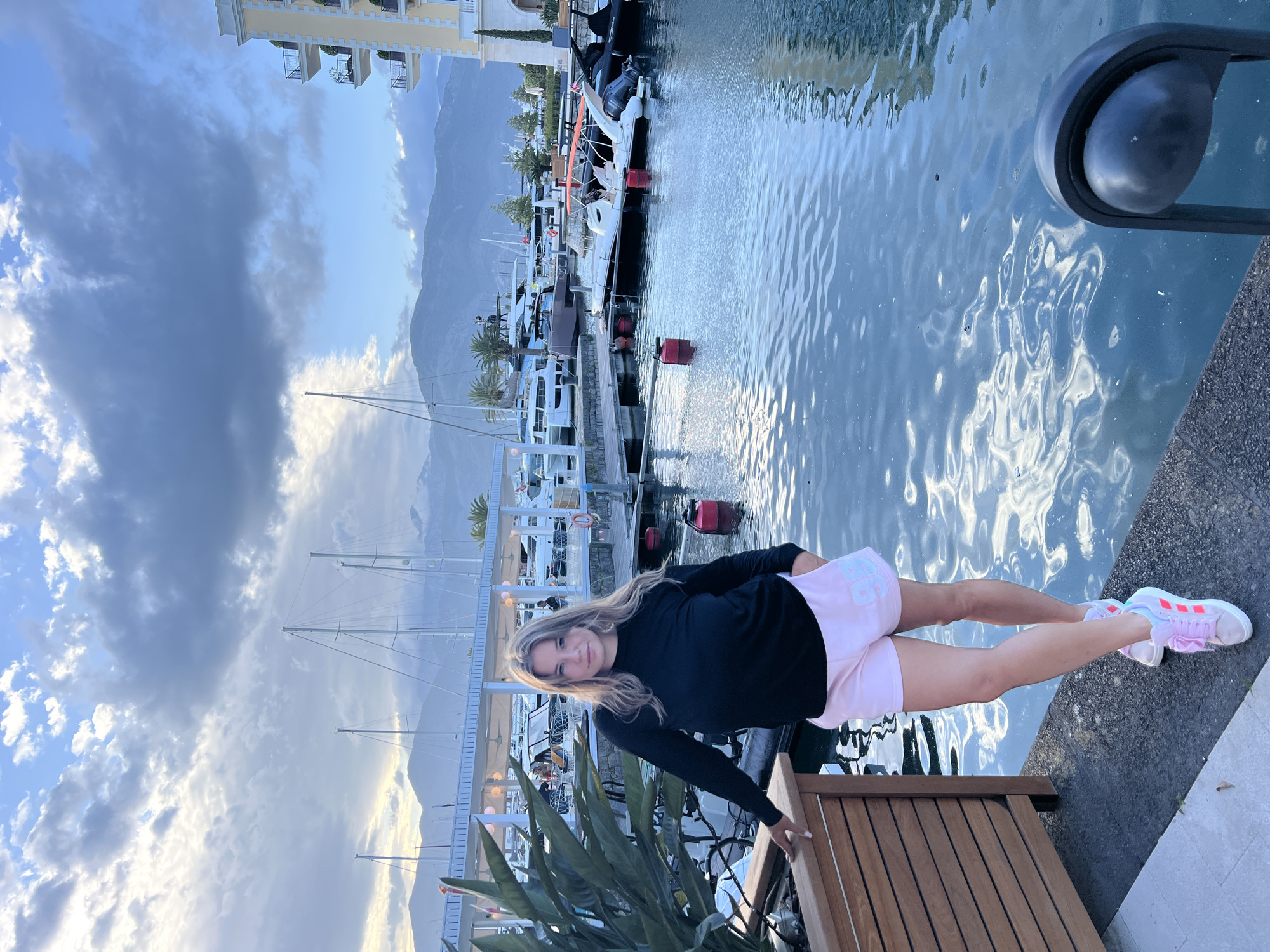 A photo shows a woman standing near a marina. The woman is leaning forward and wearing a long-sleeved black shirt, pink shorts, and pink-and-white sneakers. She has light brown hair. In the background are several boats, water, mountains, and a building. The photo was taken in Tivat, Montenegro.