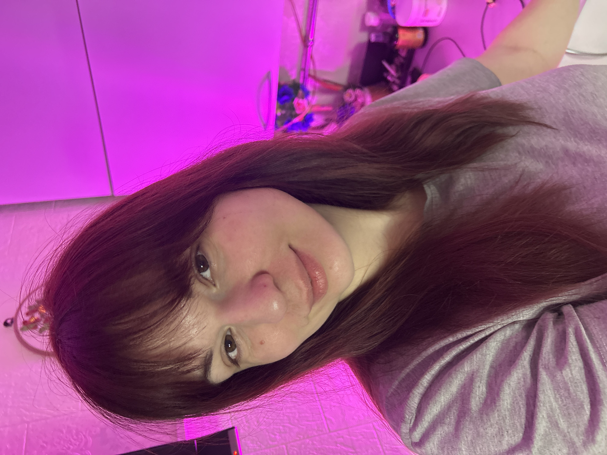 A portrait-oriented photo captures a young woman with long, dark reddish-brown hair and bangs, looking directly at the camera with a subtle smile. Her head is tilted slightly. She has light brown eyes and a fair complexion, with some natural skin texture visible. She is wearing a simple light gray t-shirt.
The setting is an indoor space, bathed in an intense magenta/purple light, suggesting artificial illumination like LED grow lights or mood lighting. This strong light casts a vibrant hue across the entire scene, saturating the background elements and creating a distinct atmosphere. On the lower left of the frame, a textured wall, possibly resembling white brick, is visible and illuminated by the purple light. In the blurry background behind her right shoulder, various household items, including bottles, containers, and wires, can be faintly discerned, suggesting a casual home environment. The image gives the impression of a casual selfie or portrait taken in an unusually lit room in Фоки, Russia. There is no visible text.