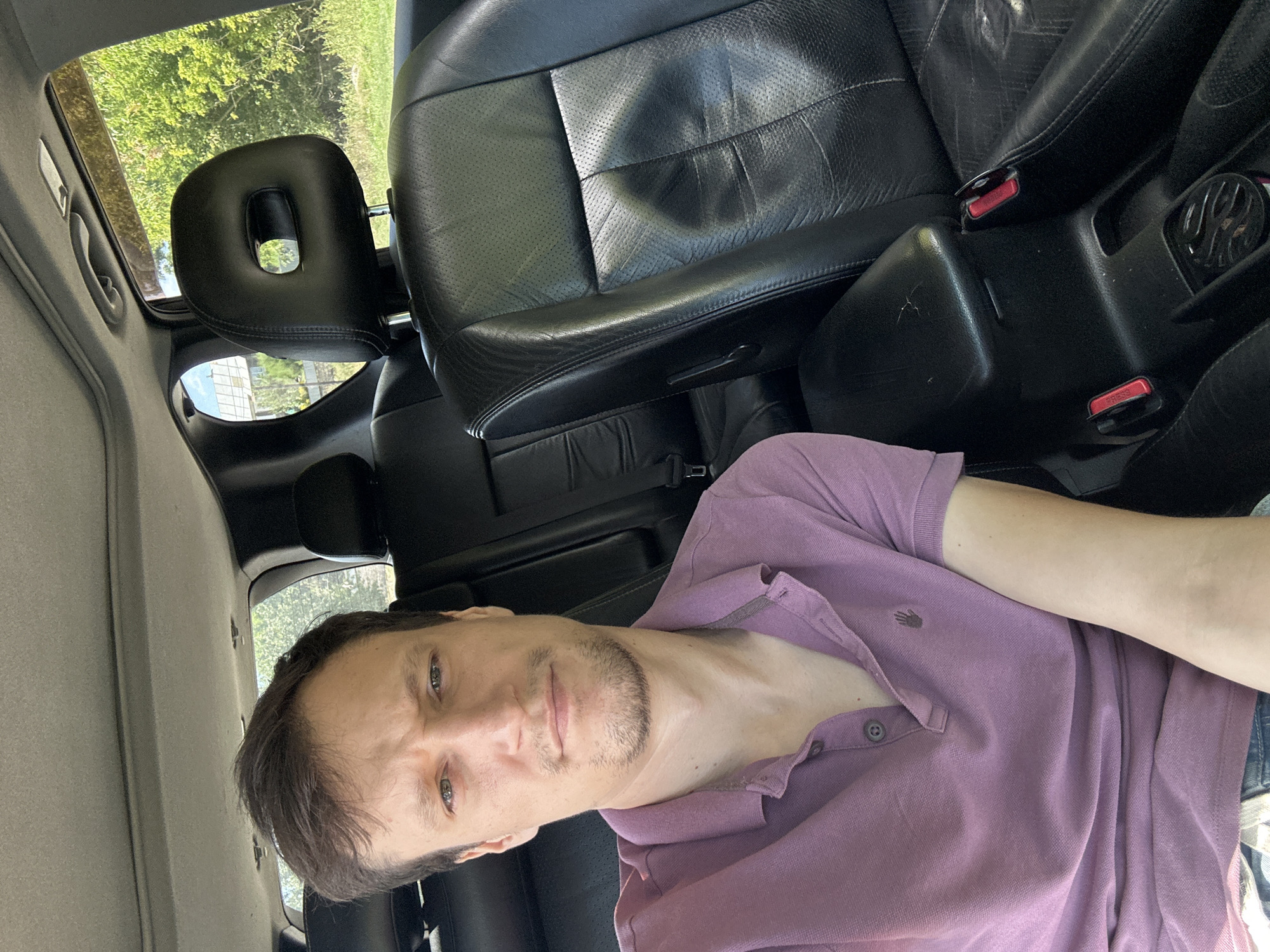 A man with short brown hair and a light stubble is taking a selfie inside a car. He is wearing a purple polo shirt. The interior of the car appears to be dark leather. Through the windows, lush green trees and a clear blue sky are visible, suggesting an outdoor, possibly rural, setting. The image was taken in Makiv, Ukraine.