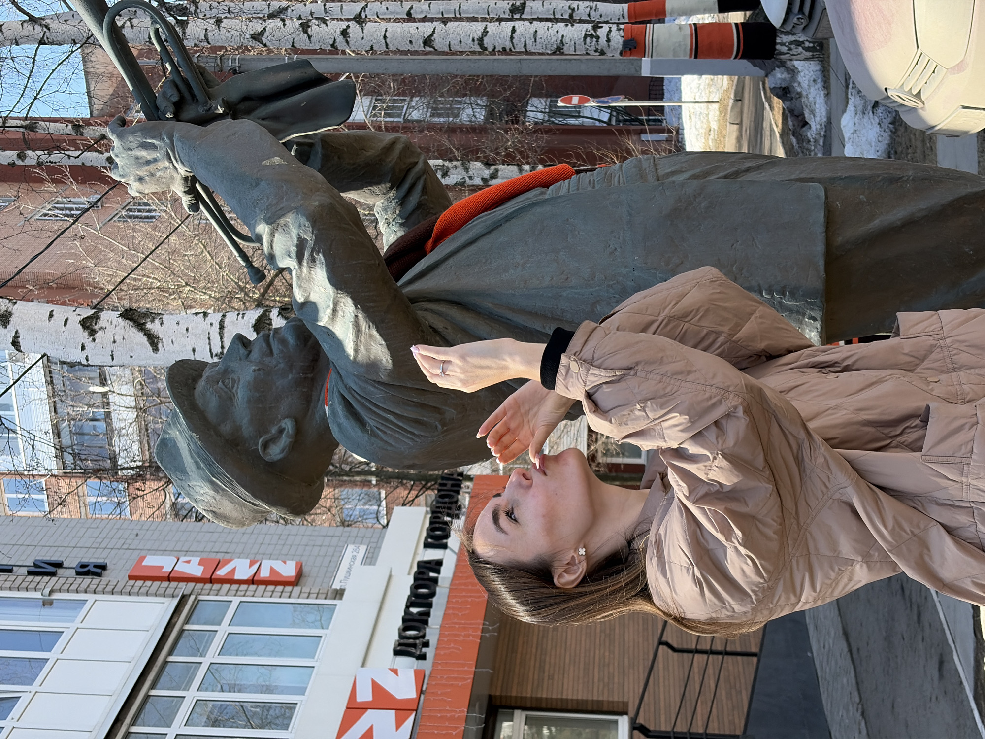 A young woman in a tan quilted jacket poses next to a statue in Izhevsk, Russia. The statue is of a man wearing a cap and holding a trumpet, and it is located outside of a building with the words "ДОКТОРА ГОРОДОНА" in Cyrillic on the building. The building is located on the street "Пушкинская". The surroundings suggest an urban environment with bare trees and snow on the ground.