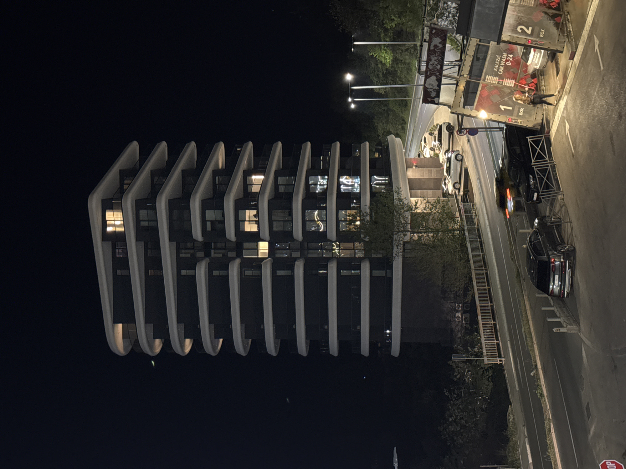 The scene is a night shot of a building, likely an apartment complex, in Boreti, Montenegro. The building is tall with a unique architectural design featuring repeating curved, white structures along its facade. The building has multiple floors, with some windows showing lights from within.
Next to the building, on the right side of the image, there's a road with several parked cars. A car wash facility, labeled "KALEZIC CAR WASH 0-24", is visible, with a person standing nearby holding what appears to be a mop. A "STOP" sign is positioned on the street's edge. Street lights are present along the road. The environment is mostly dark due to nighttime. Trees and some vegetation are also visible.