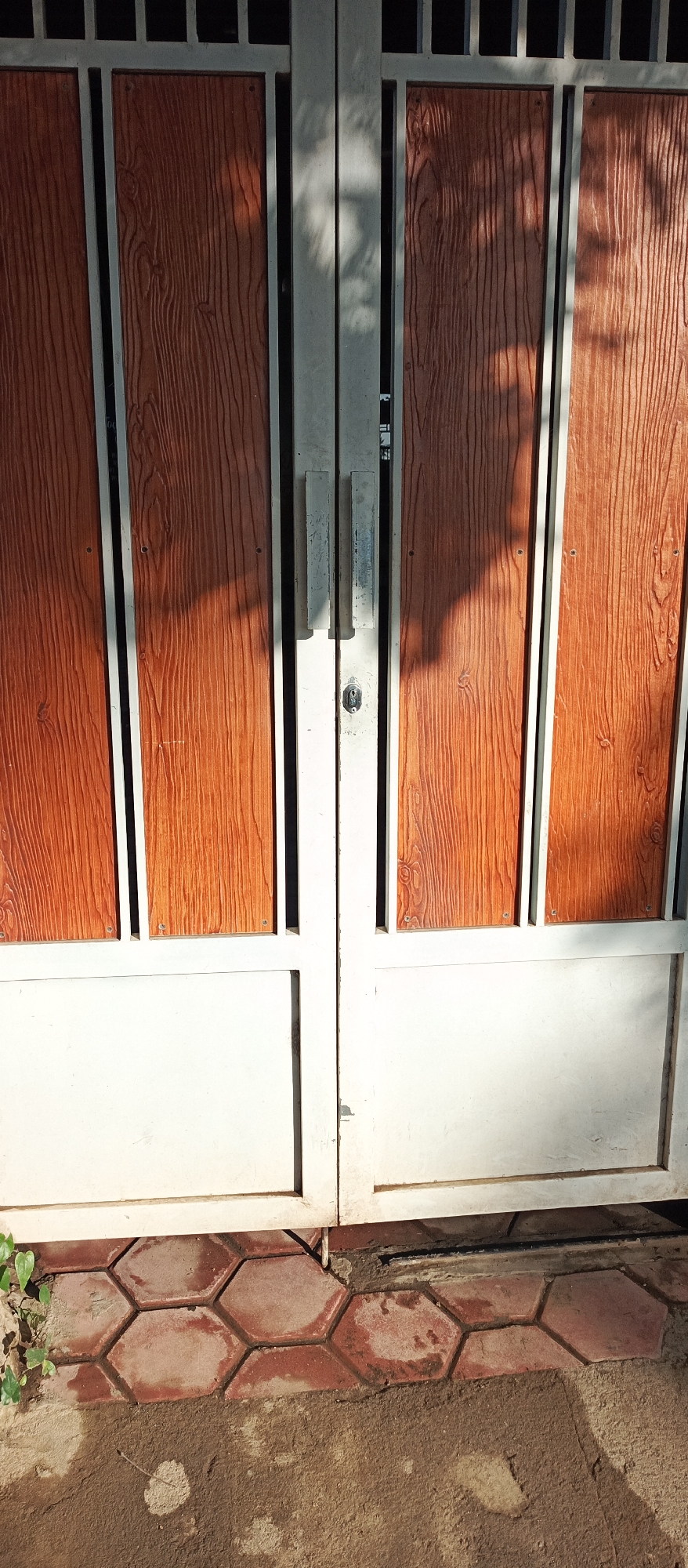 This is a medium-angle, eye-level, close-up shot of a pair of closed, light-colored metal gates in front of a residential property in Arjasa, Indonesia. The gates are divided into sections, with the top sections featuring a reddish-brown material with a visible wood grain pattern, framed by a silver metal border. The bottom sections of the gates are a solid, light-colored metal. A silver door handle is visible on the right gate, and a keyhole is centered on the seam between the two gates. Sunlight casts dappled shadows on the gates, suggesting the presence of overhead foliage. The ground in front of the gates is paved with reddish-brown hexagonal tiles, some of which appear worn and stained. The edge of the pavement meets a dirt and concrete area, with a small amount of green foliage visible on the left side of the frame. The overall impression is one of a functional, outdoor entrance to a home in a tropical or subtropical climate.