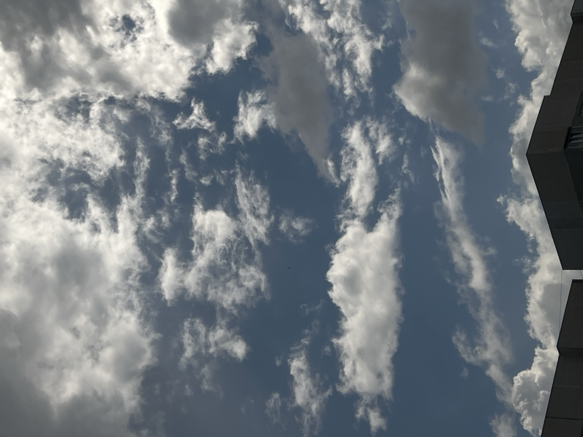 The image shows a cloudy sky over the rooftops of buildings in Ulyanovsk, Russia. The clouds are a mix of white and grey, with some appearing dark and heavy, suggesting a possibility of rain. The sky is a deep blue, peeking through the cloud cover. The buildings are modern, with flat roofs and grey facades. The perspective is looking upwards, emphasizing the vastness of the sky. There are no people or animals visible in the scene. The overall atmosphere is one of a typical overcast day.