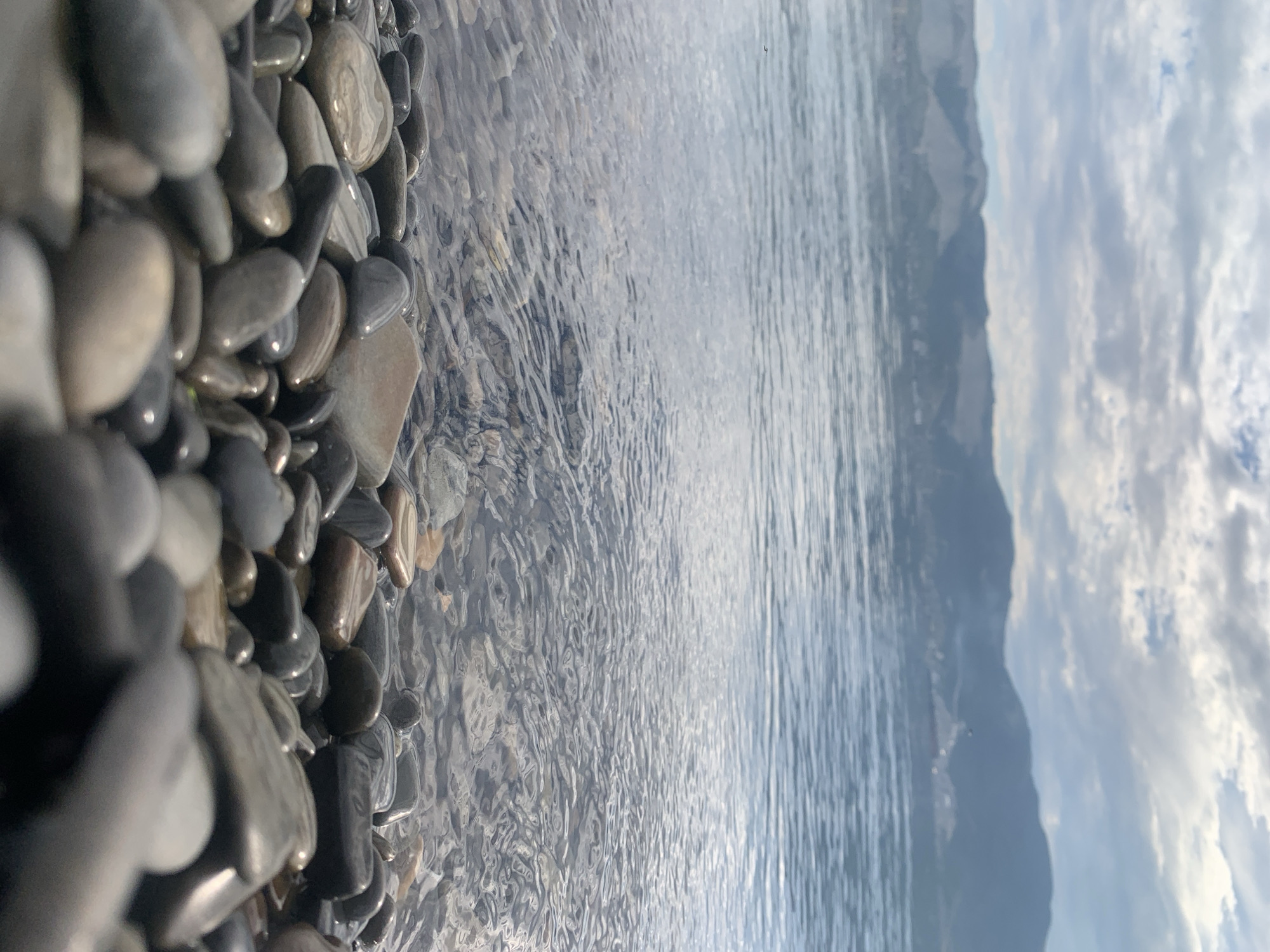The image depicts a low-angle view of a pebble beach along a body of water, likely the sea, with a mountainous landscape in the background under a cloudy sky. The foreground is dominated by smooth, rounded pebbles of various sizes and colors, ranging from dark gray to lighter beige and some with reddish-brown hues. The water's edge is visible, with clear, shallow water revealing more pebbles beneath the surface, rippled by gentle waves. In the distance, hazy mountains form a backdrop, with a thin line of what appears to be settlement or structures visible along their base. The sky is filled with dynamic clouds, suggesting a daytime scene with diffused sunlight. There are no people or man-made structures immediately evident on the beach itself.
The image appears to be taken from the shoreline of Novorossiysk, Russia, a Black Sea port city known for its coastline, mountainous terrain, and pebble beaches. The specific location would likely be one of the numerous beaches that line the city's coastal area. The presence of mountains in the background is characteristic of the landscape surrounding Novorossiysk.