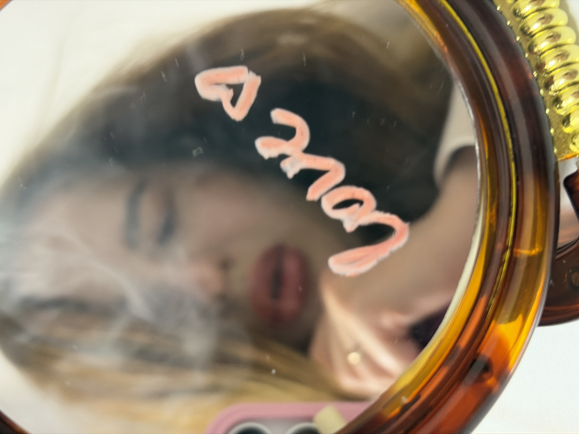 A close-up, reflective shot captures a person's blurred face in a round mirror with a tortoiseshell-patterned frame, possibly in Krasnodar, Russia. The word "love" with a heart symbol is written in a light, peachy-pink substance, resembling lipstick or a creamy cosmetic, directly onto the mirror's surface. The person's lips, visible in the reflection, are pursed, suggesting a kissy face or an expression associated with affection. Their long, light brown hair frames their face, which is largely out of focus.
On the right side of the mirror, a decorative golden, ridged element is visible, possibly part of the mirror's stand or an ornate handle. Below the reflection, a hint of a pink object, perhaps a phone case or a makeup item, can be seen. The background outside the mirror's frame is a plain, light-colored surface. The overall impression is one of intimacy and playfulness, possibly a self-portrait conveying affection or self-love.